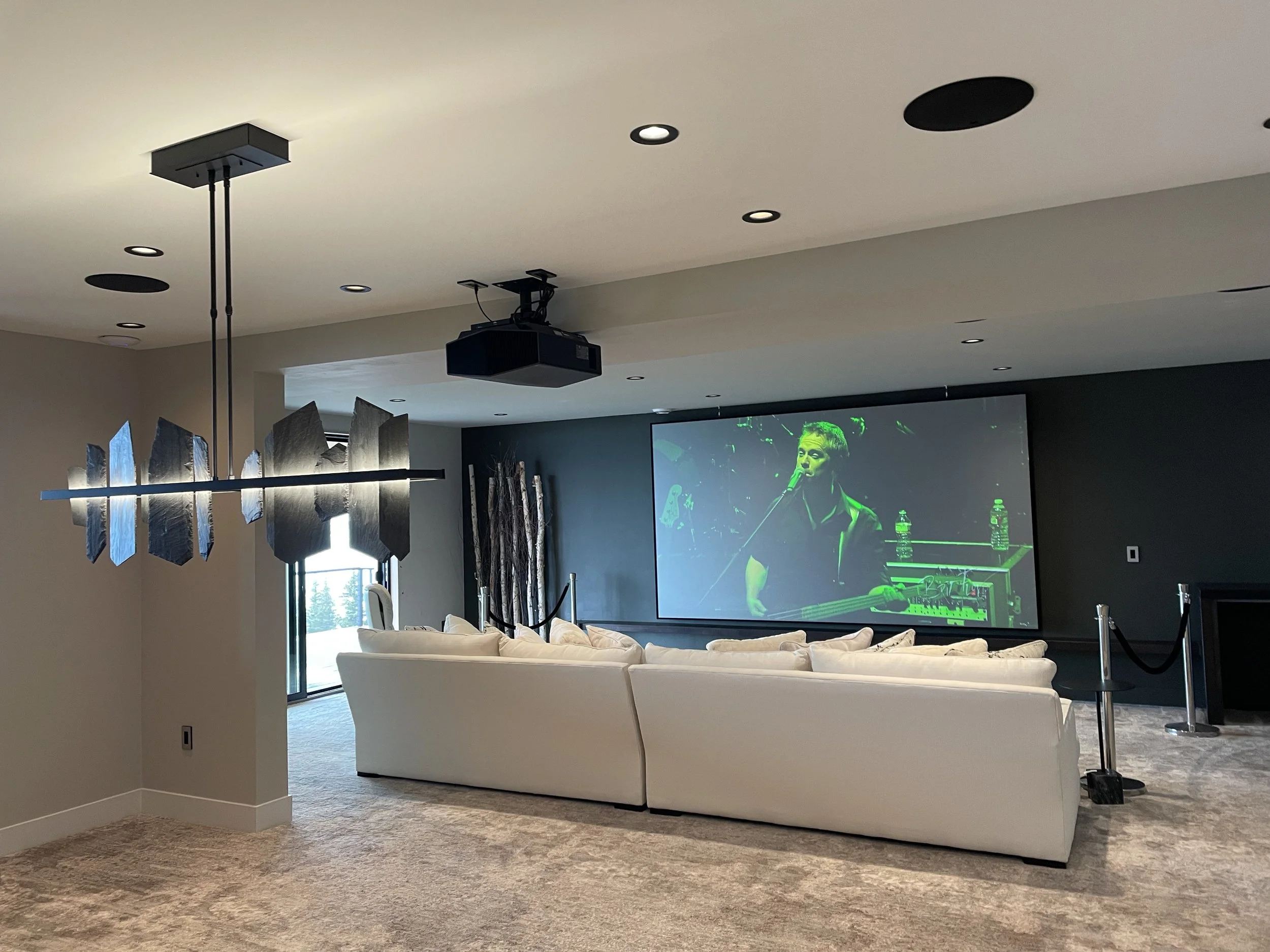 Living room with a large Screen Innovations projection screen showing a man playing guitar, white sofa, modern chandelier, carpeted floor, black roped barrier, and a window with blinds. A Sony 4K HDR Laser Projector is seen mounted to the ceiling.