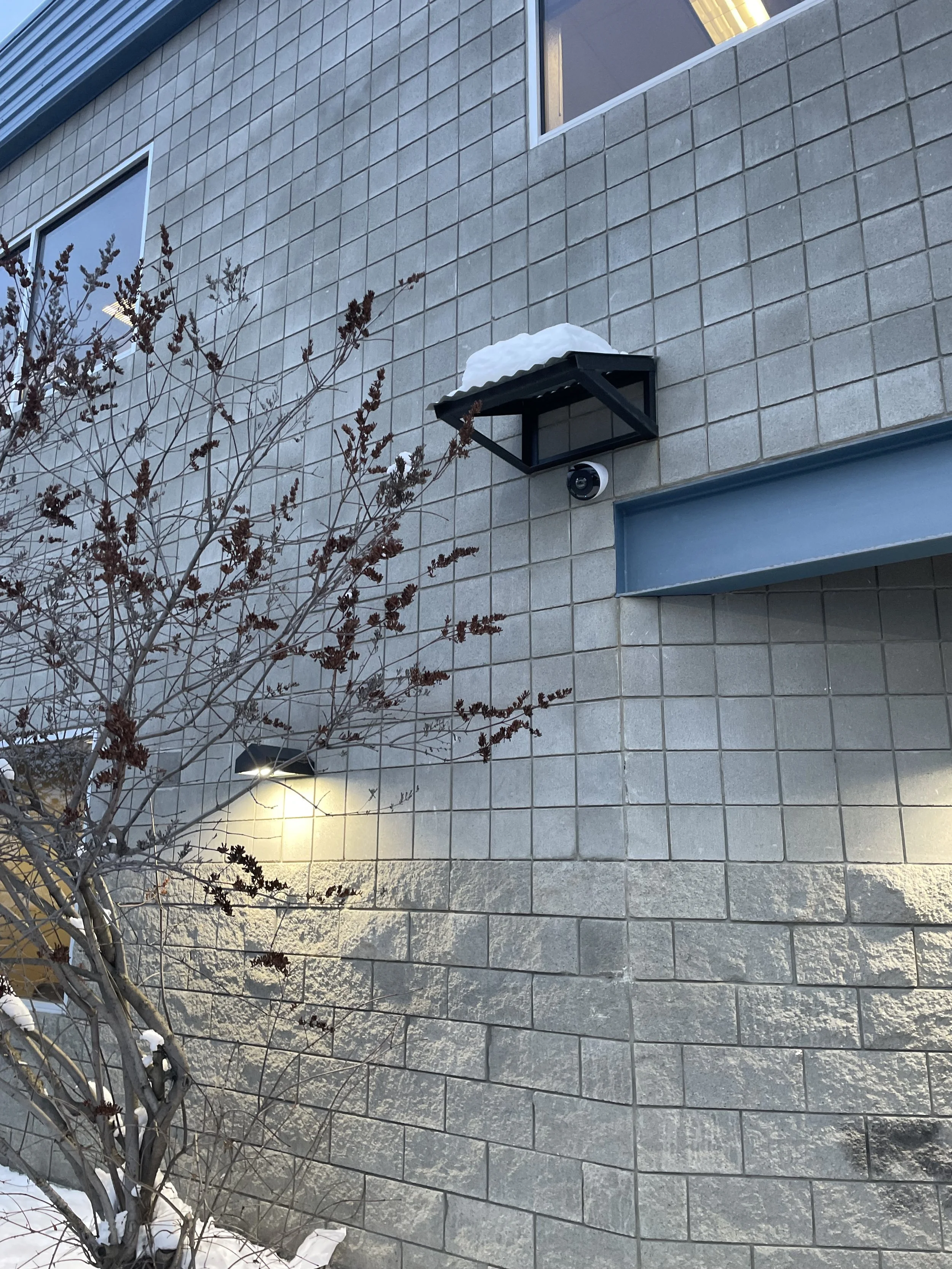 Exterior wall of a building with beige tiles, a window, a security camera, snow on a small metal roof, a light fixture illuminating a leafless tree, and patches of snow on the ground.