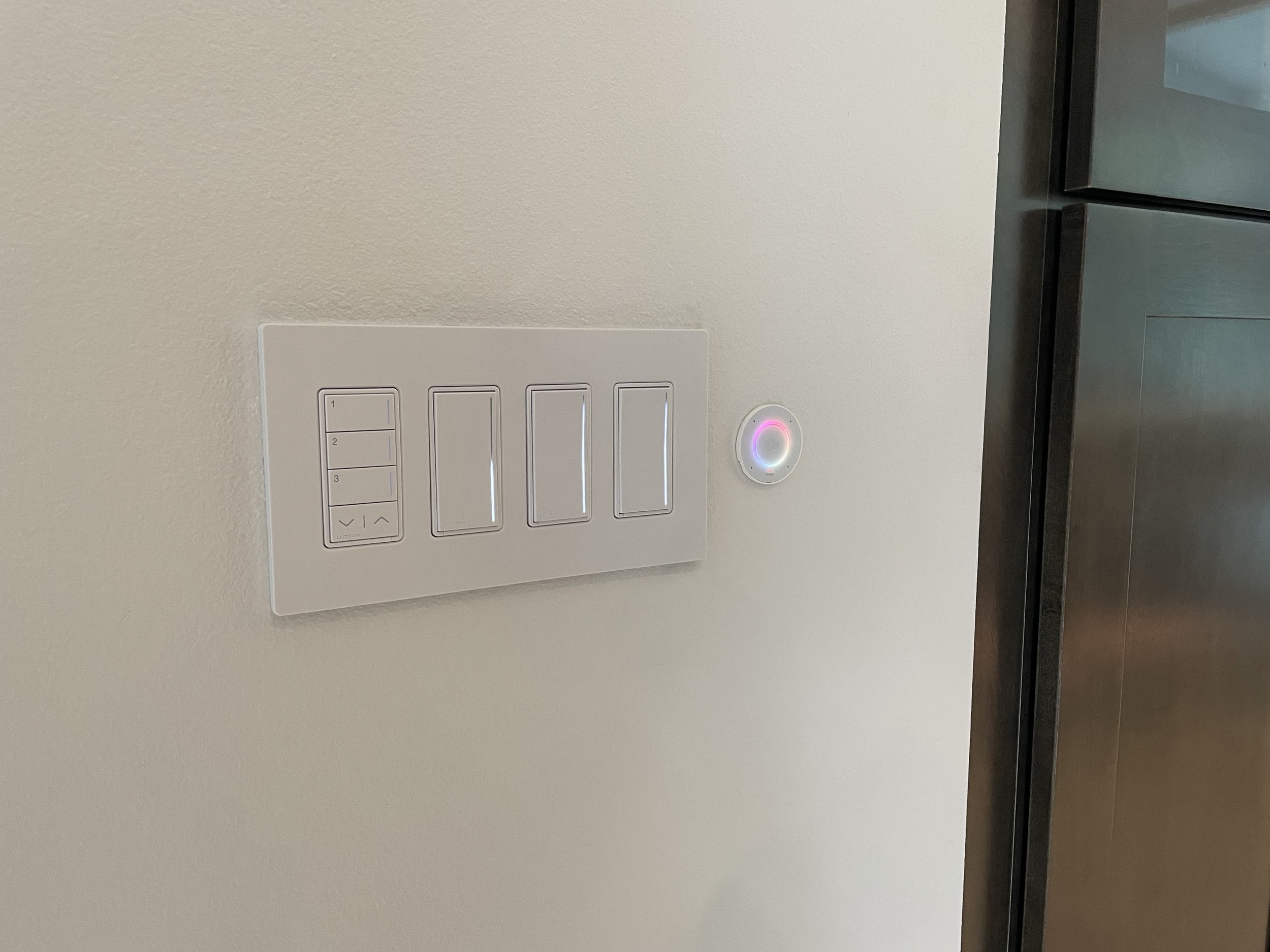 A white wall with a control panel with four switches and a circular device with a purple light next to it.