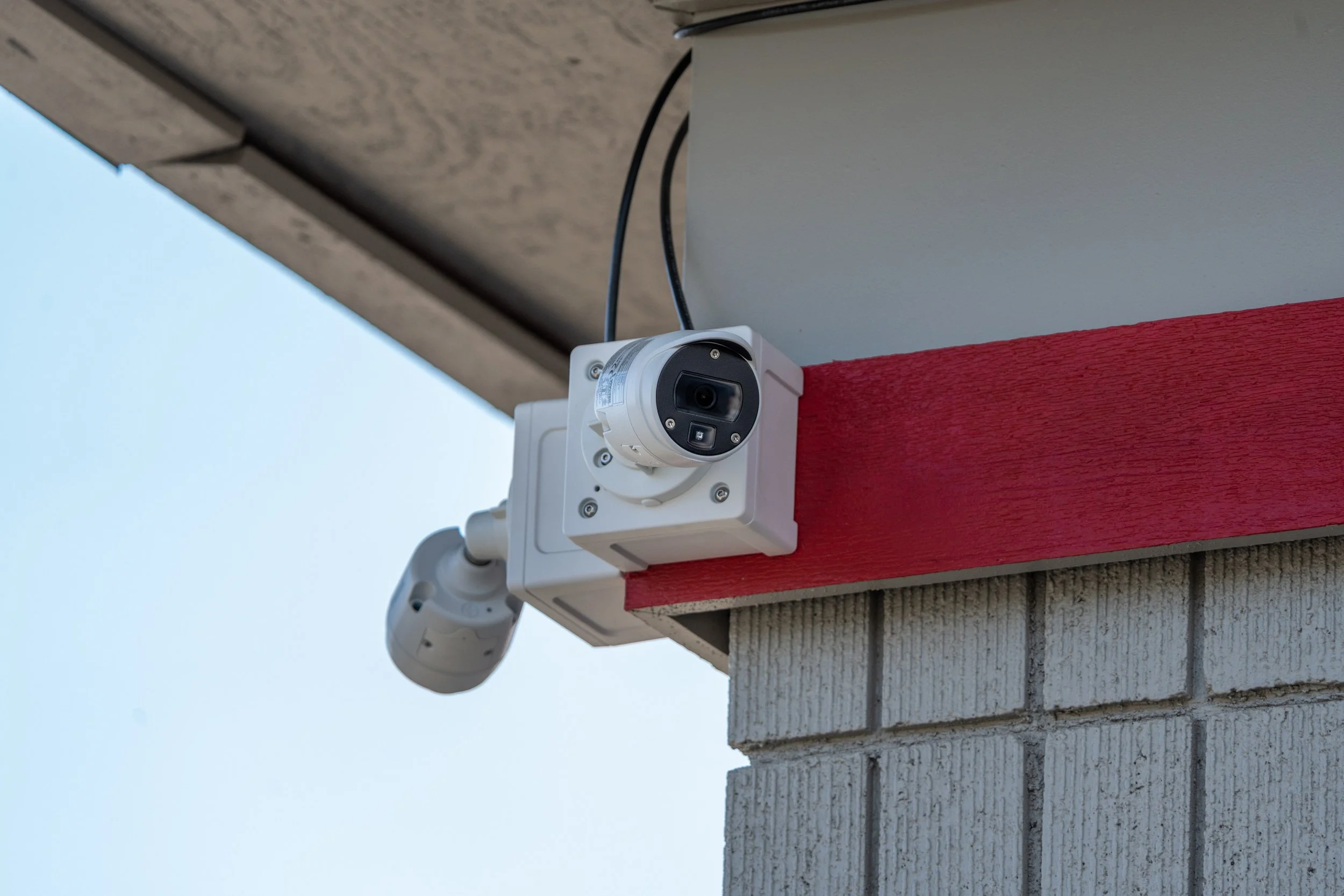 A security camera mounted on the corner of a building wall under the roofline, with a clear sky in the background.