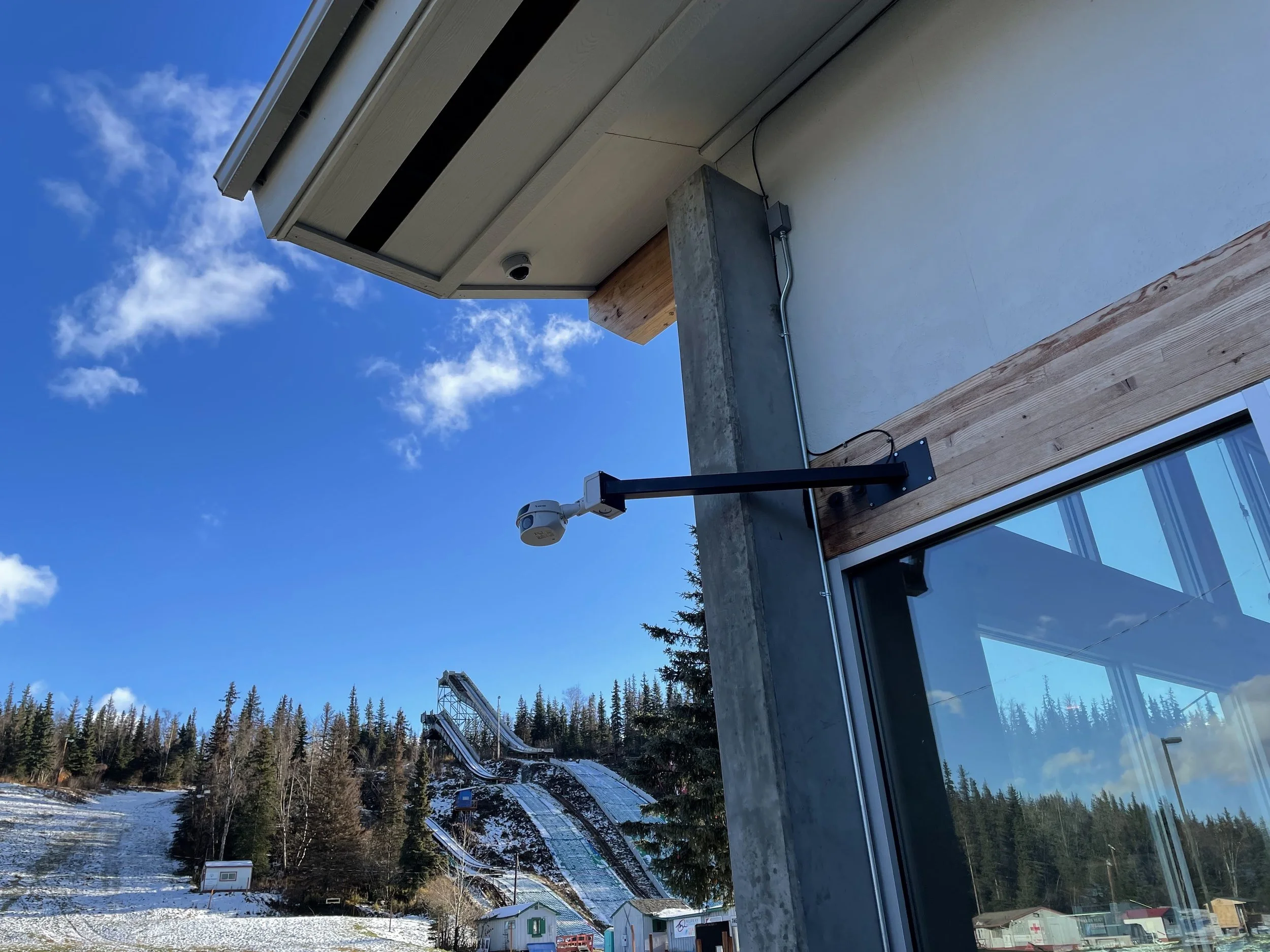 A security camera mounted on a black arm attached to a building with a glass window and wooden siding, overlooking a snowy landscape with ski jumps in the background.