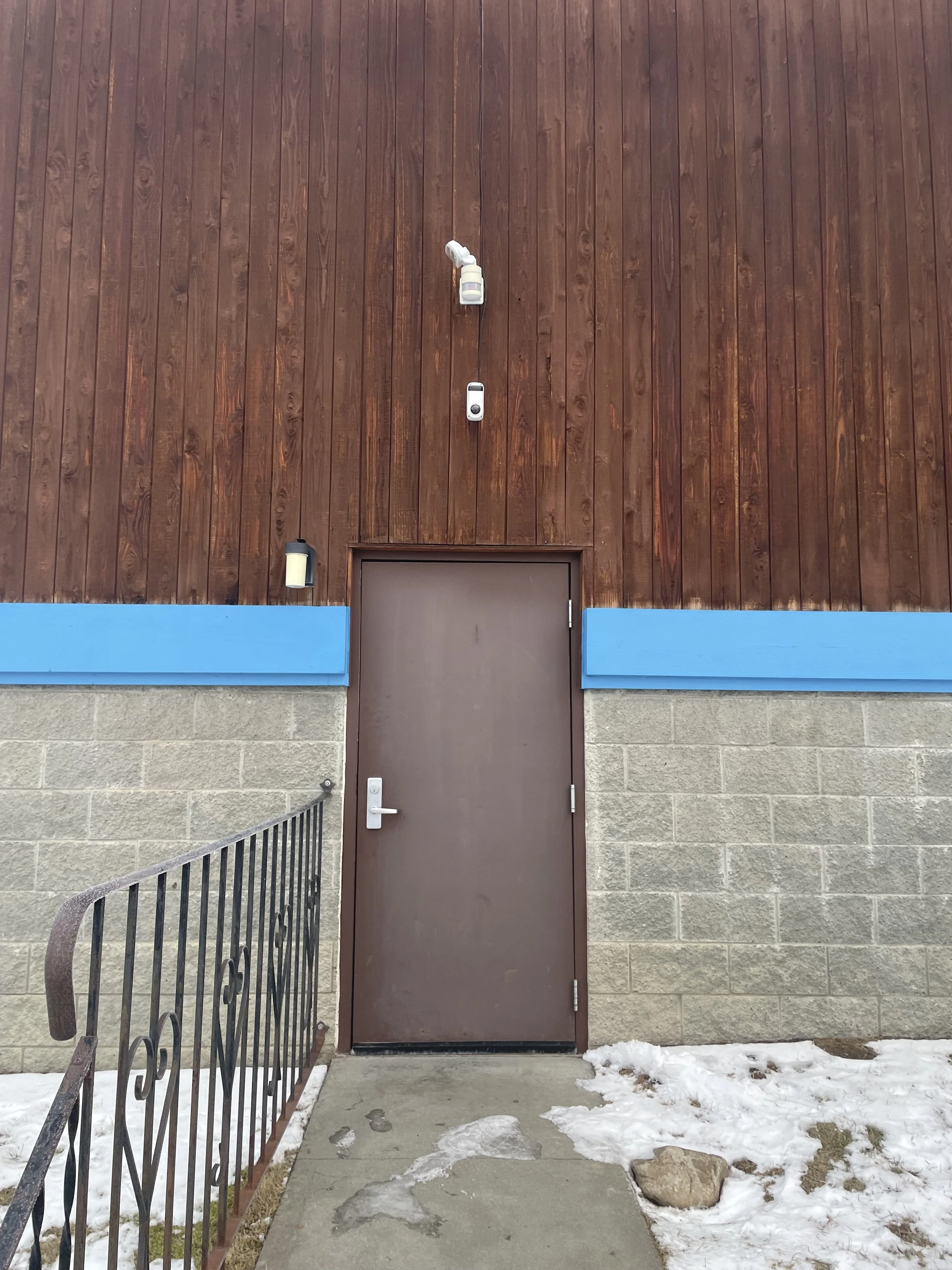 A brown door on a building with wood paneling and blue trim, snow on the ground, and a black metal railing on the left.