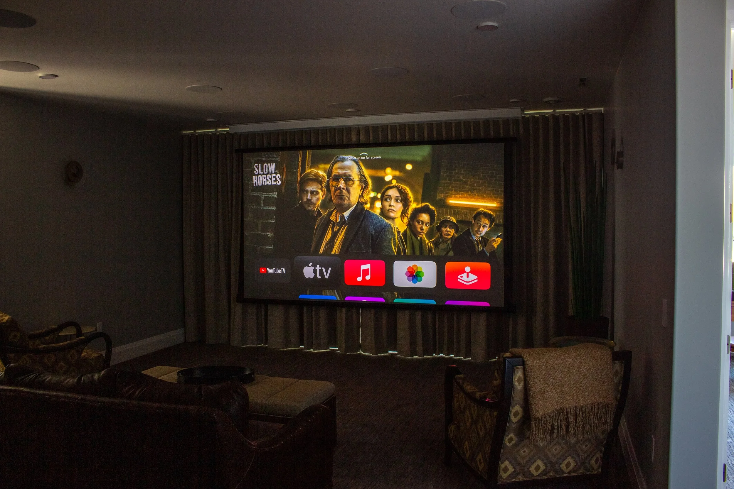 A home theater room with a large screen showing scene from the show 'Slow Horses'. The screen displays Netflix icons for YouTube TV, Apple TV, Apple Music, Photos, and Arcade at the bottom. There are couches and chairs facing the screen, with curtain