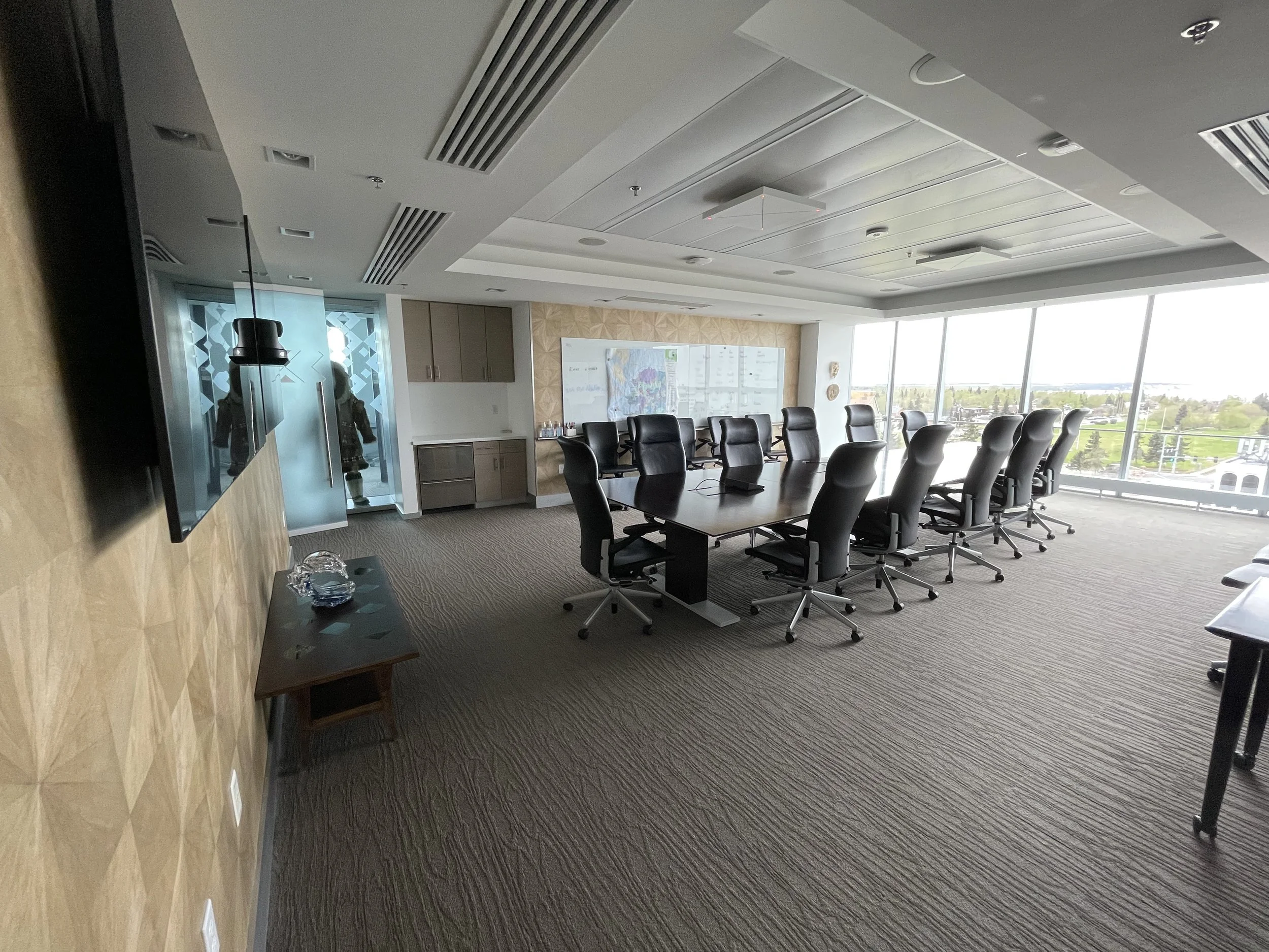 Empty modern conference room with a long table, black swivel chairs, large window with city view, wall-mounted TV, and a whiteboard.