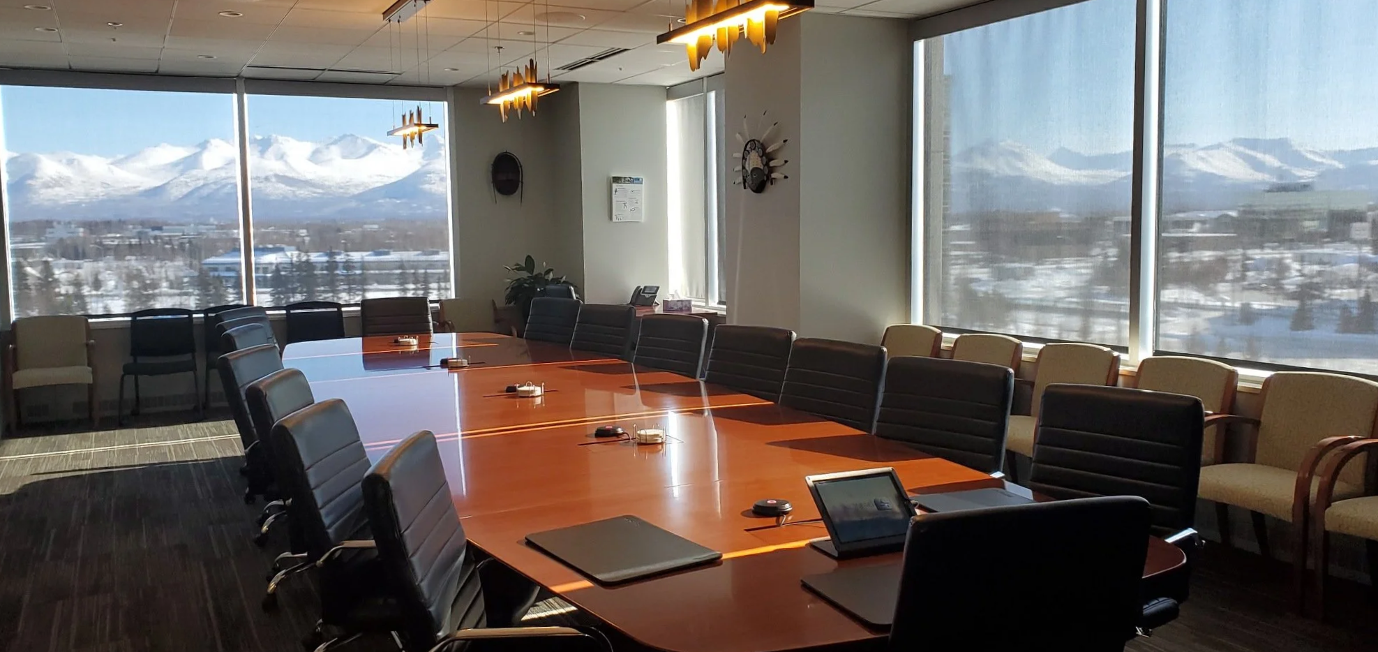 Empty conference room with a large oval wooden table, black and beige chairs, some with armrests, and large windows showing a snowy mountain landscape background.
