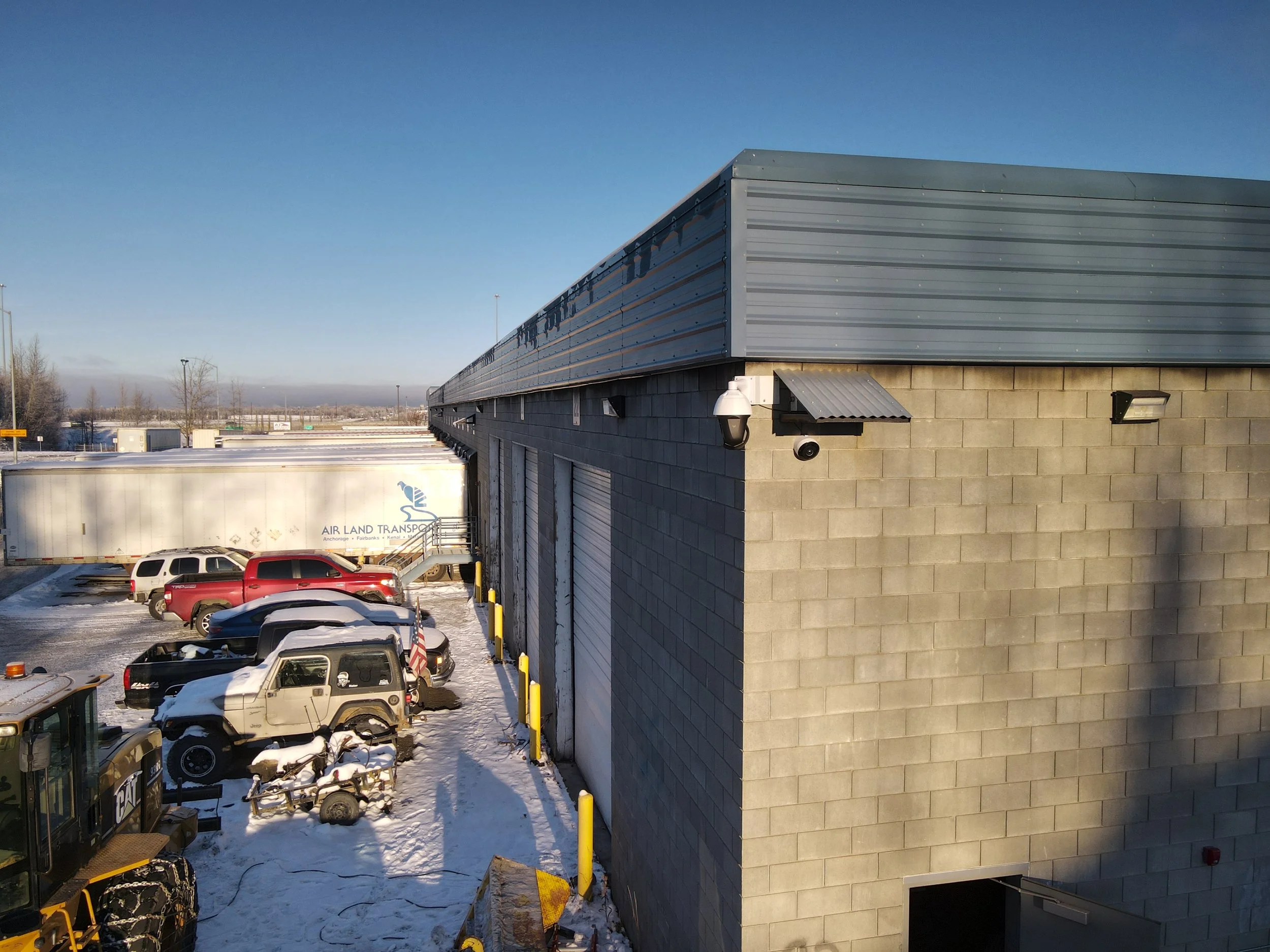 Parking lot with snow, several parked cars, a tractor, and a large industrial building with a brick wall and metal siding, under a clear blue sky.
