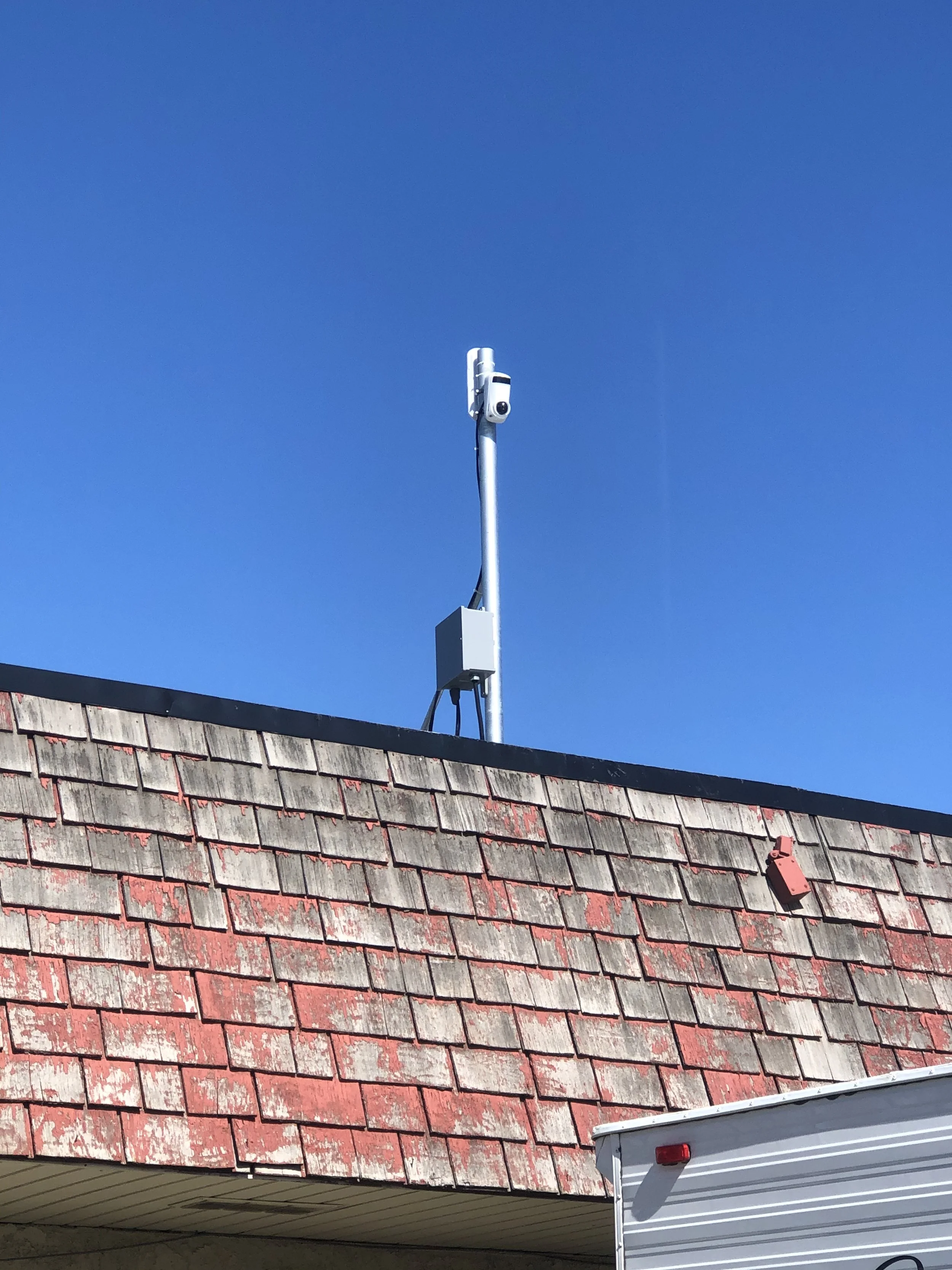 A close-up of a roof showing weathered red shingles and a security camera mounted on a pole against a clear blue sky.