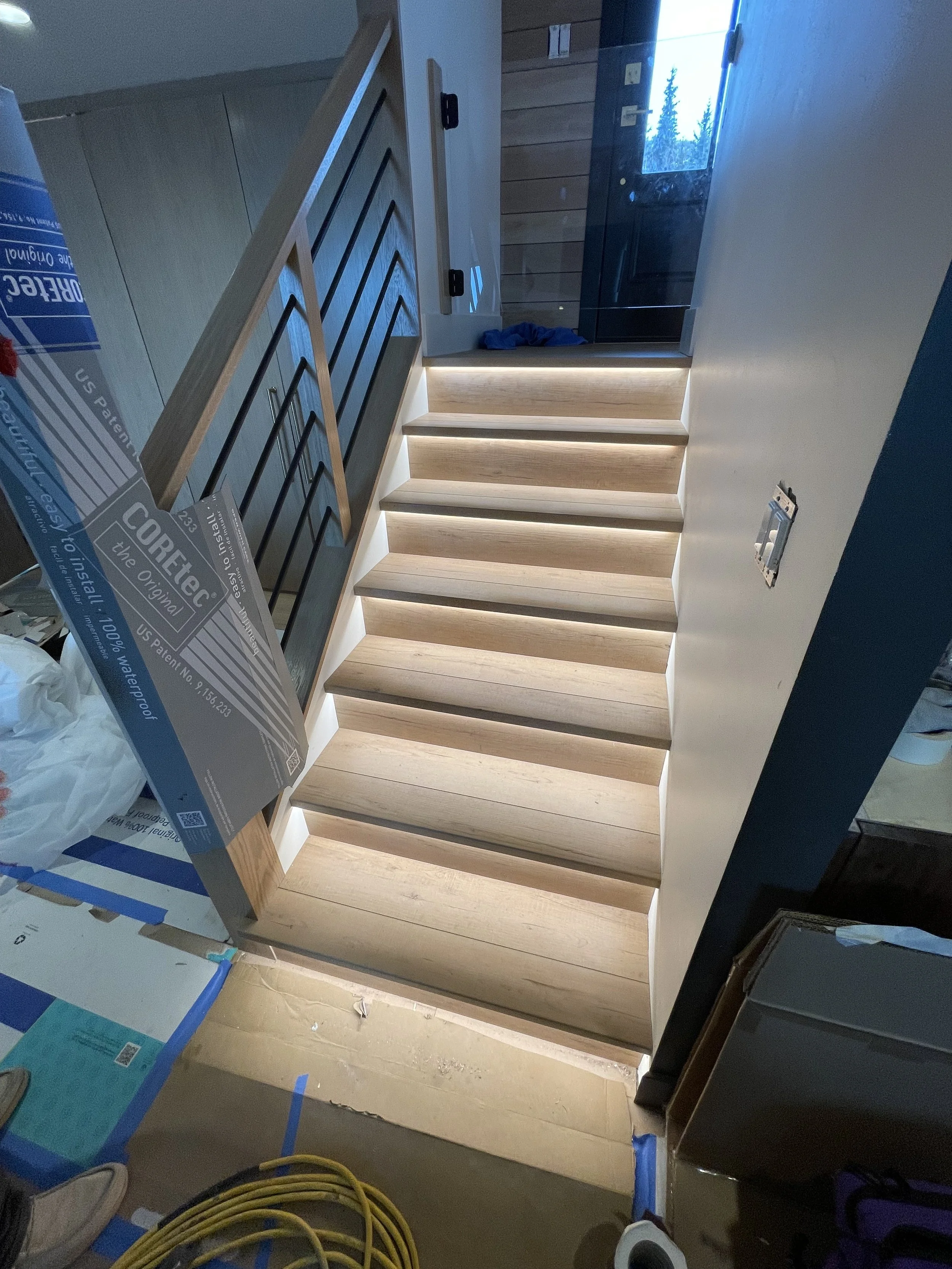 Modern staircase with wooden steps and LED lighting underneath, leading to a glass door at the top with a view outside.