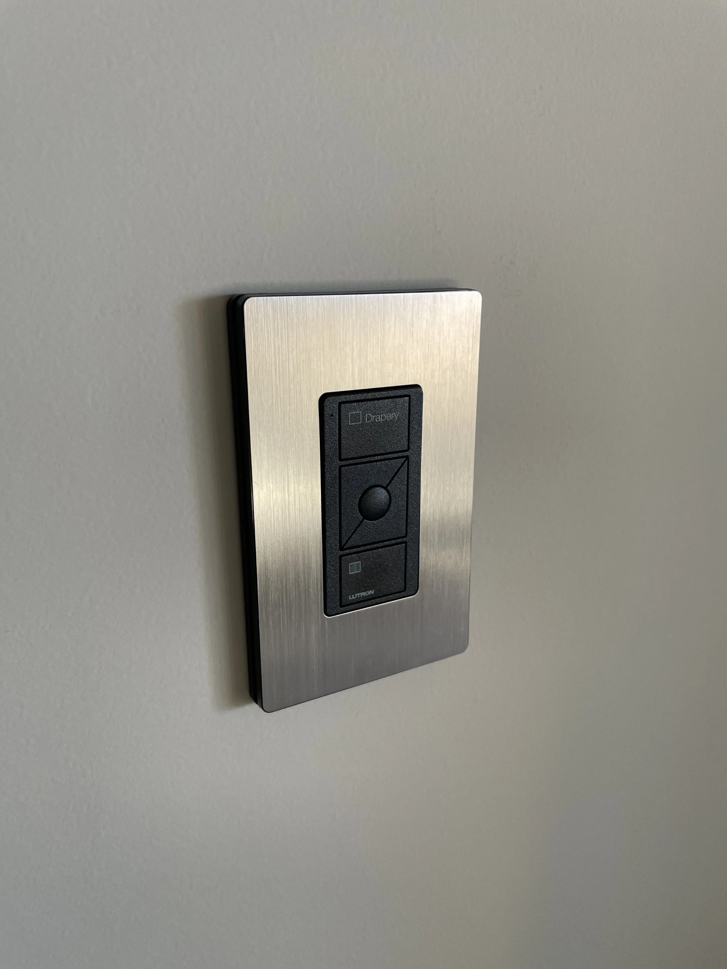 A brushed stainless steel Lutron faceplate containing custom shade controls, mounted on a plain wall.