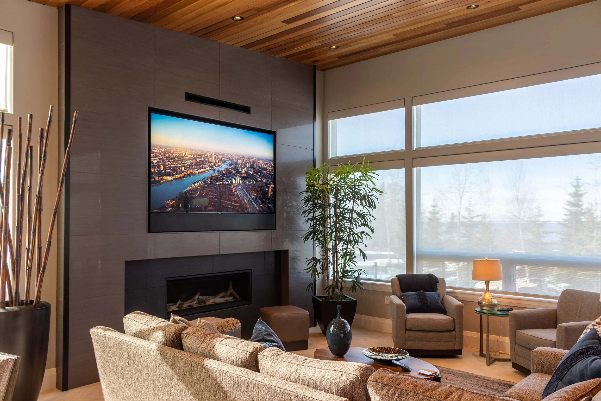 Modern living room with a wall-mounted TV, a fireplace below it, a beige sofa, two armchairs with pillows, a side table with a lamp, and large windows with city and outdoor views.