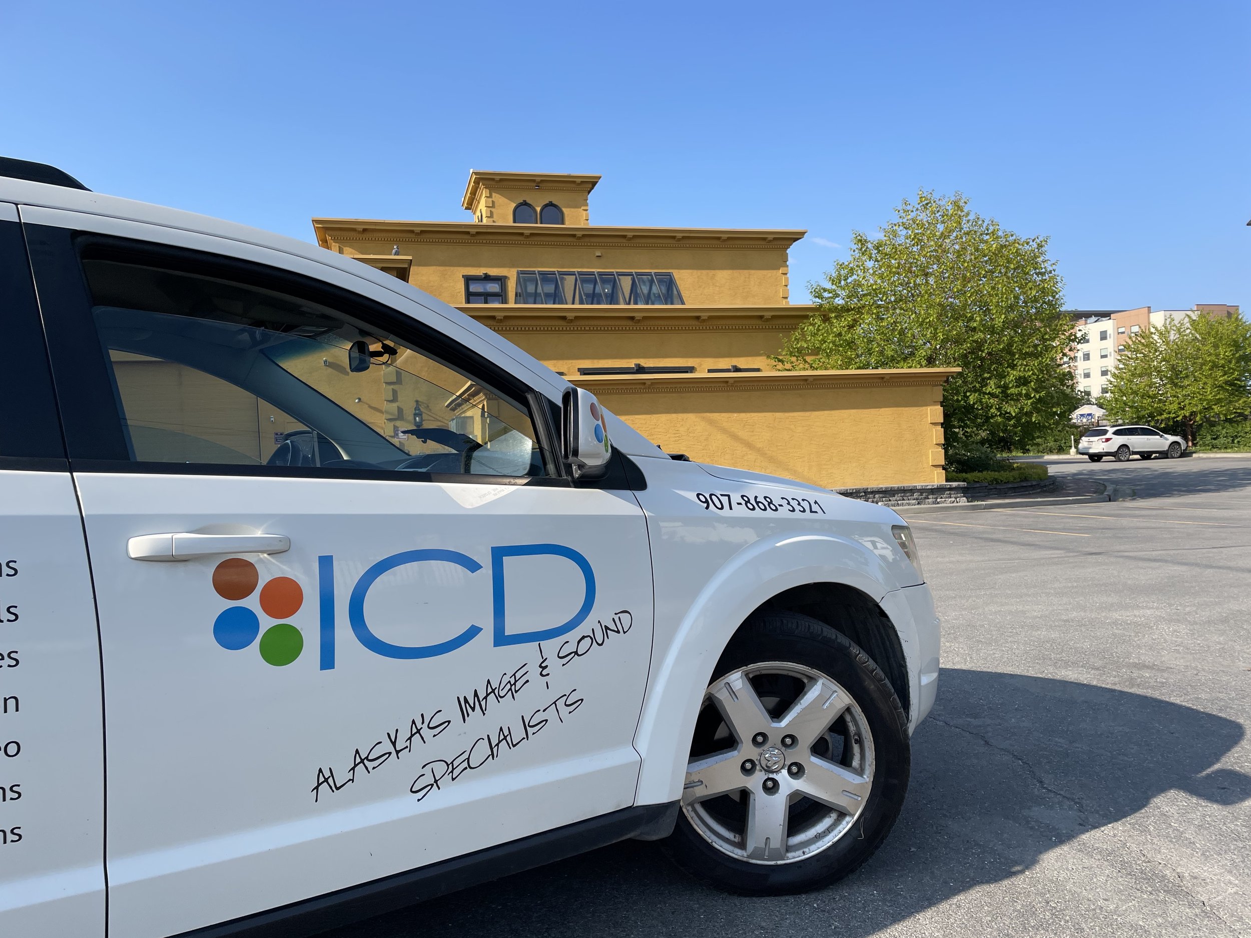 A white vehicle with the logo and text 'ICD Alaska's Image & Sound Specialists' parked in an outdoor parking lot near a yellow building with trees and another car in the background.