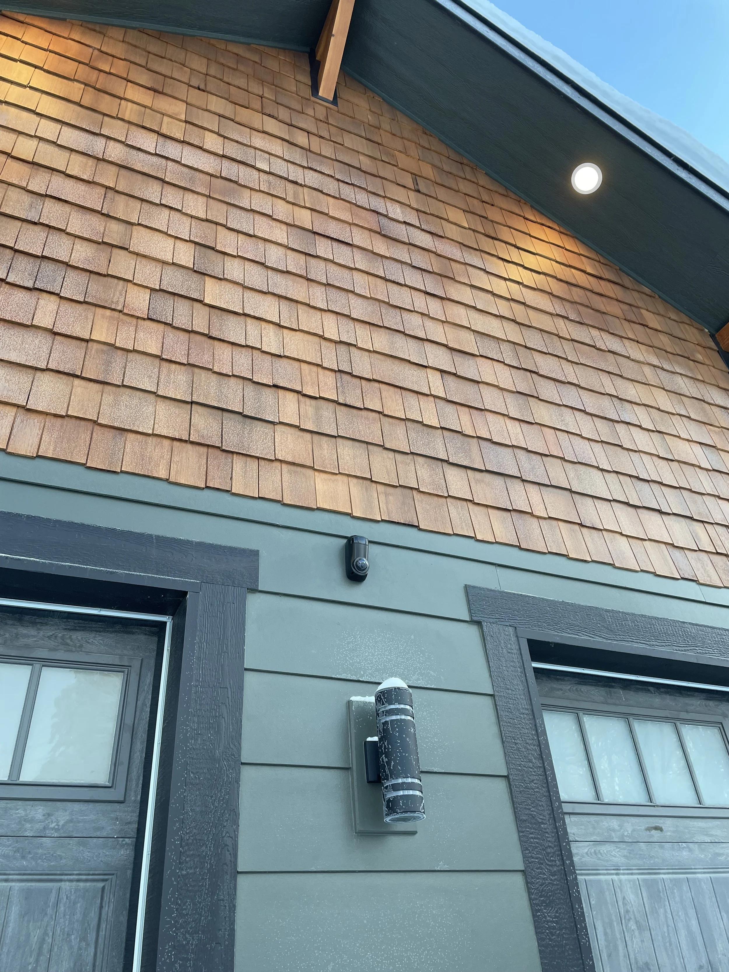 Exterior view of a house showing a roof with brown shingles, a greenish wall with a black security camera, a cylindrical outdoor light fixture, and a window with a dark frame and decorative trim.
