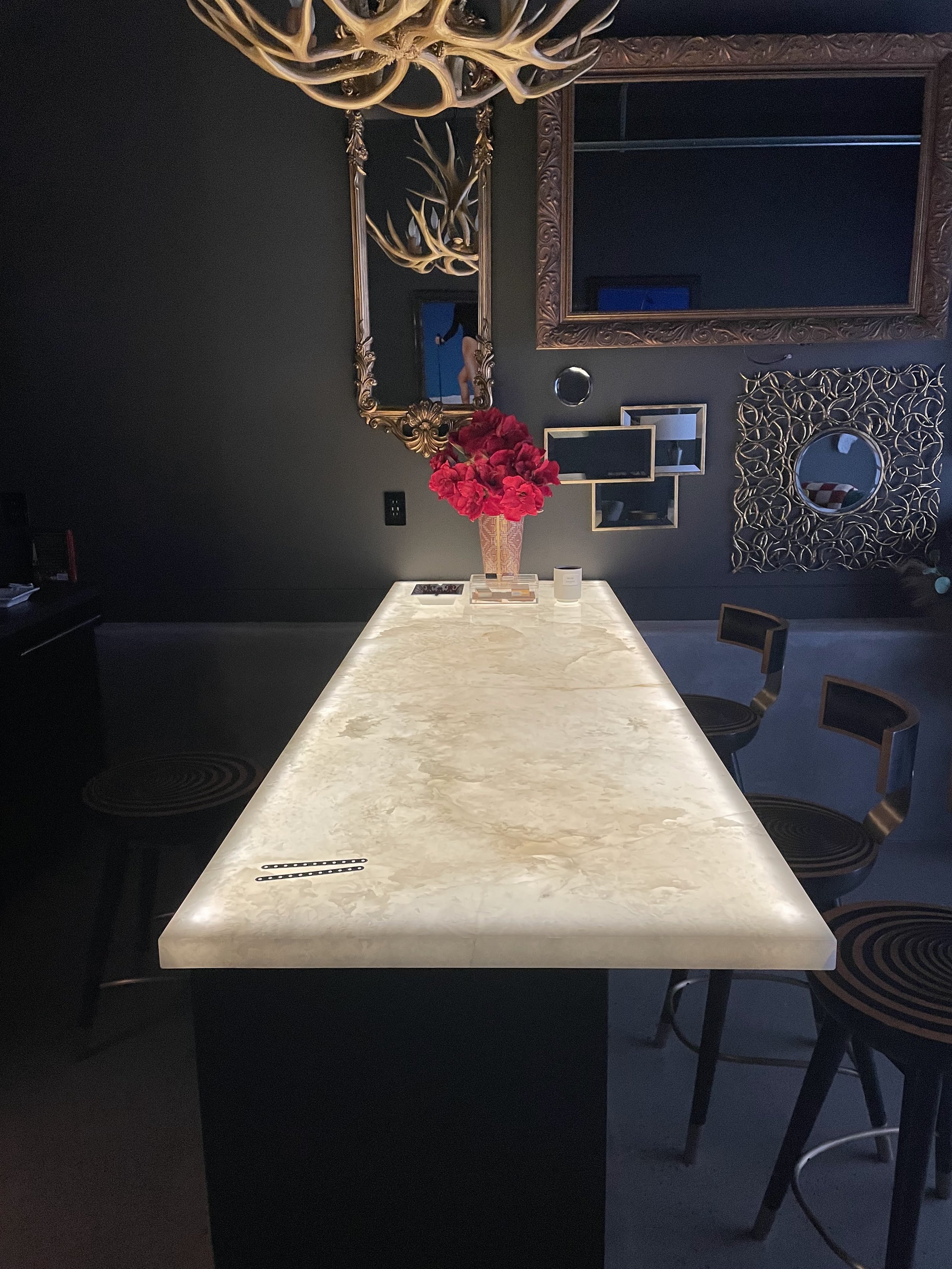 A modern dining room with a glowing marble table, decorative wall mirrors, framed art, a bouquet of red flowers, and gold antler chandeliers.