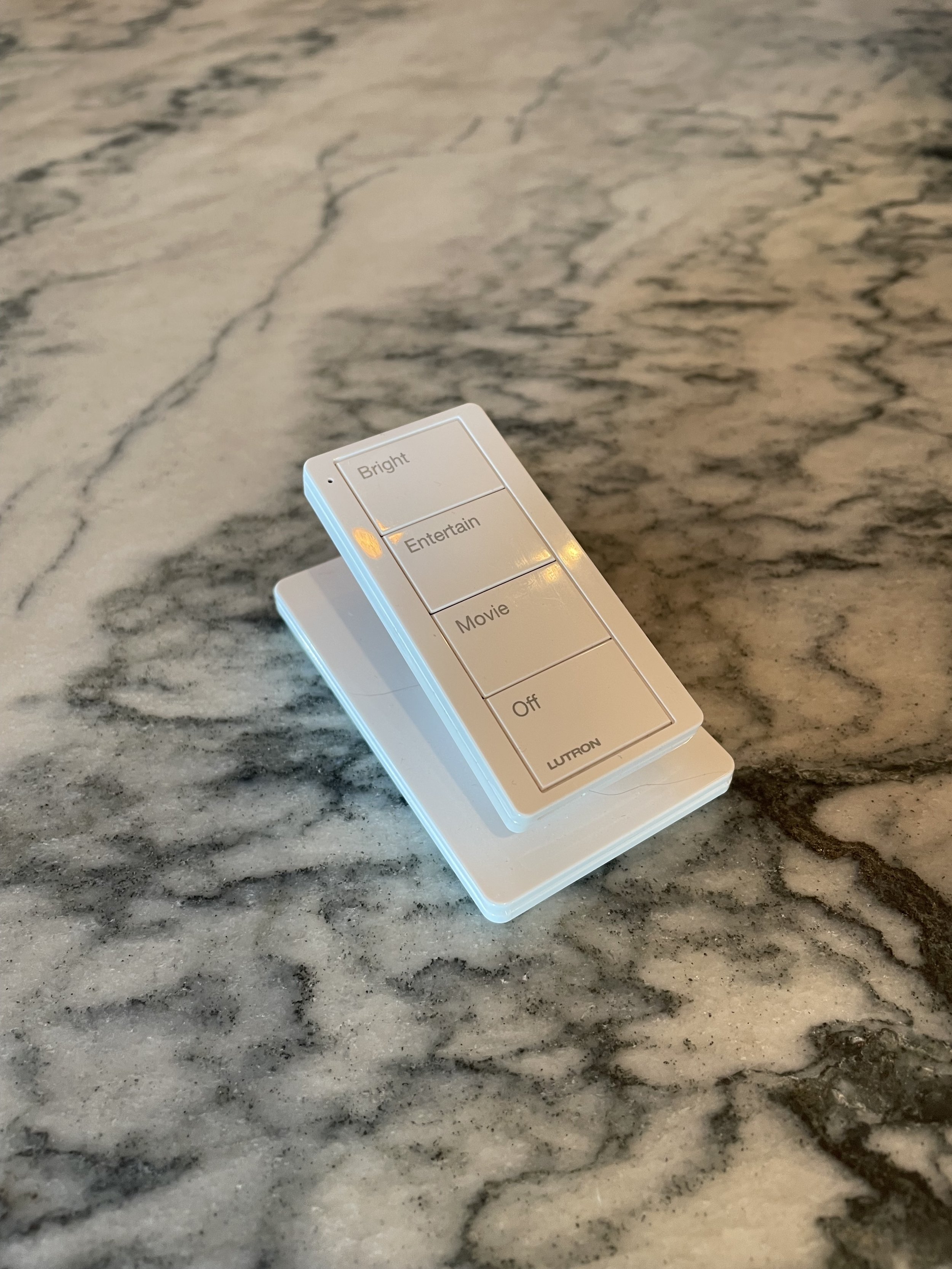 A Lutron Pico remote control with custom buttons labeled Bright, Entertain, Movie, Off, and Lutron, resting on a marble surface.