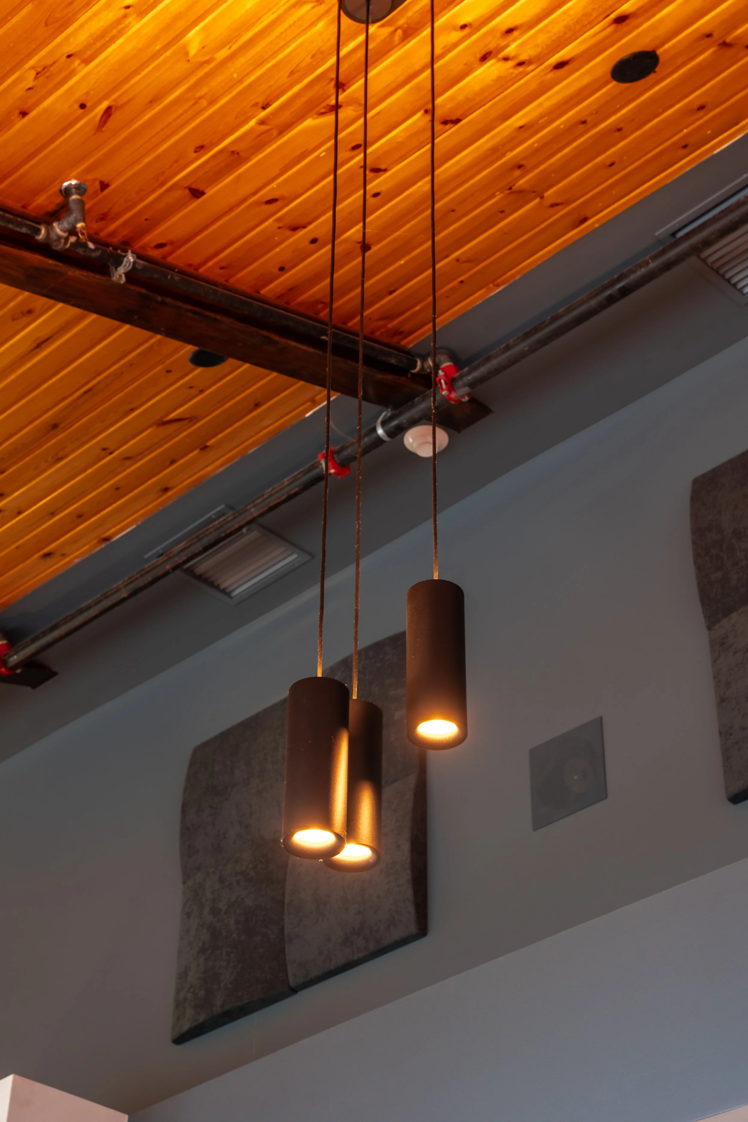 Three cylindrical pendant lights hanging from a ceiling with wood paneling and visible pipes.
