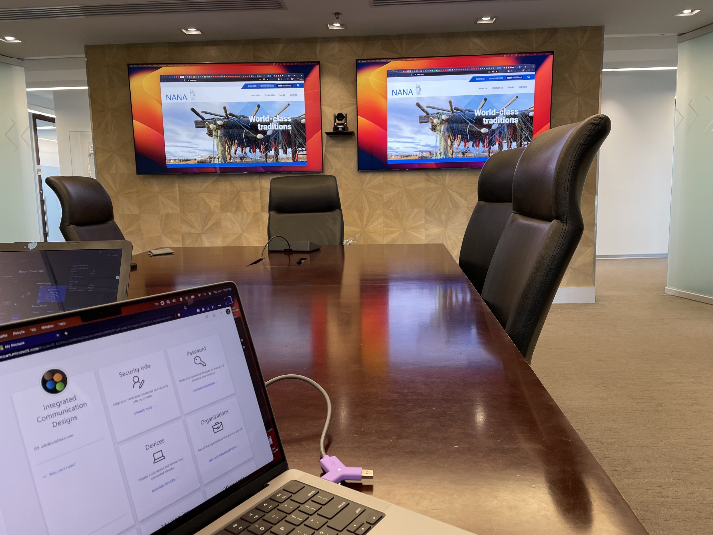 A conference room with a large wooden table and six black leather chairs. Two large flat-screen monitors on the wall display a website called 'NANA' with the slogan 'World-class traditions'. A laptop is open on the table, showing a webpage titled 'In