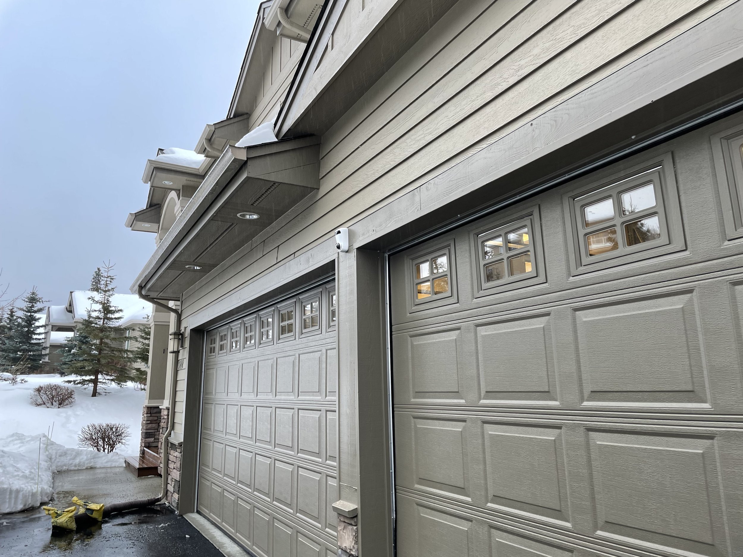 Close-up of a beige garage door with small window panes at the top, part of a beige house with horizontal siding, snow on the roof, and a snow-covered yard with trees in the background.