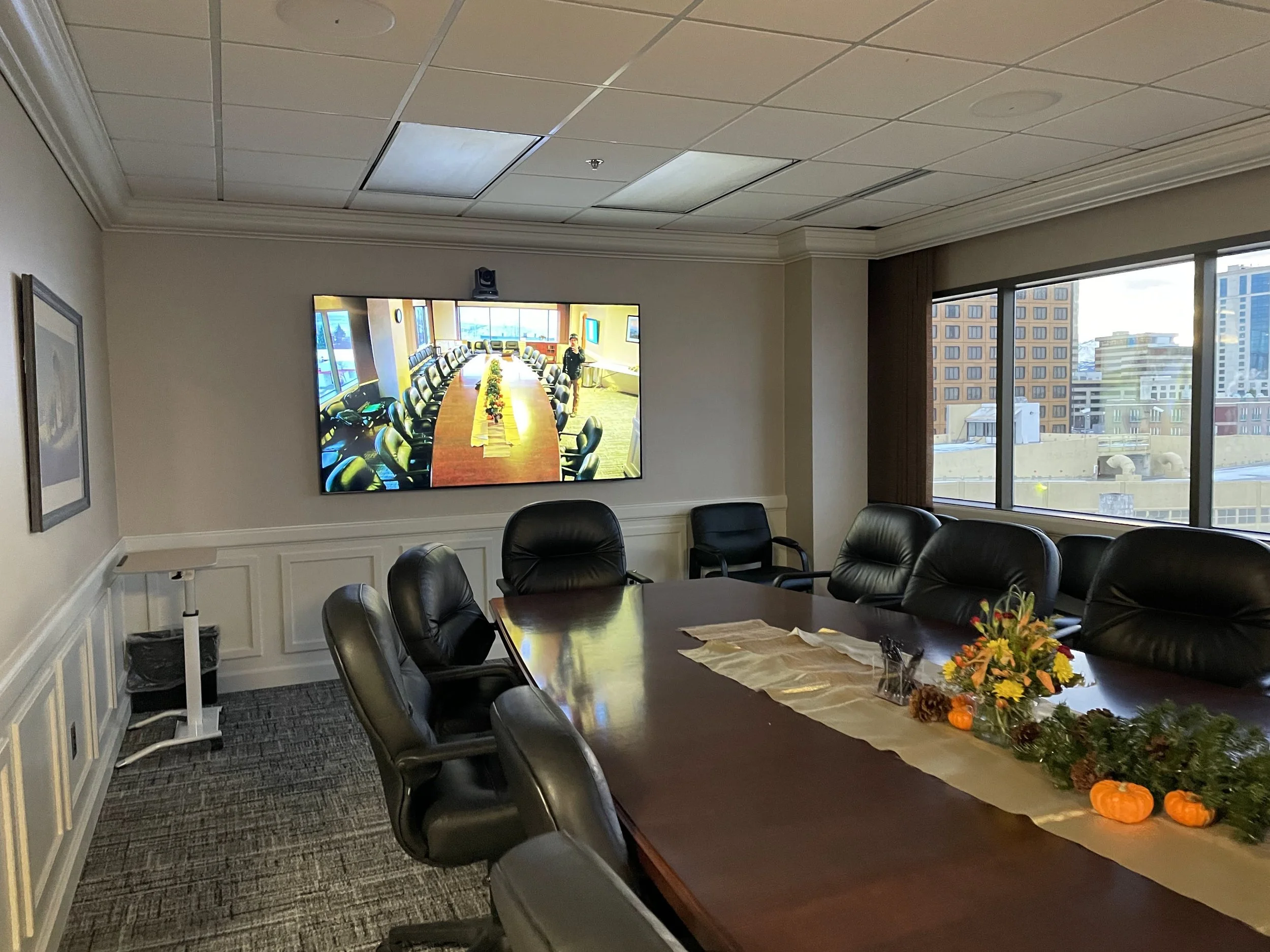 Empty conference room with a large table, black leather chairs, flowers and small pumpkins as decorations, a wall-mounted TV showing a different room with a long conference table, window with city view.
