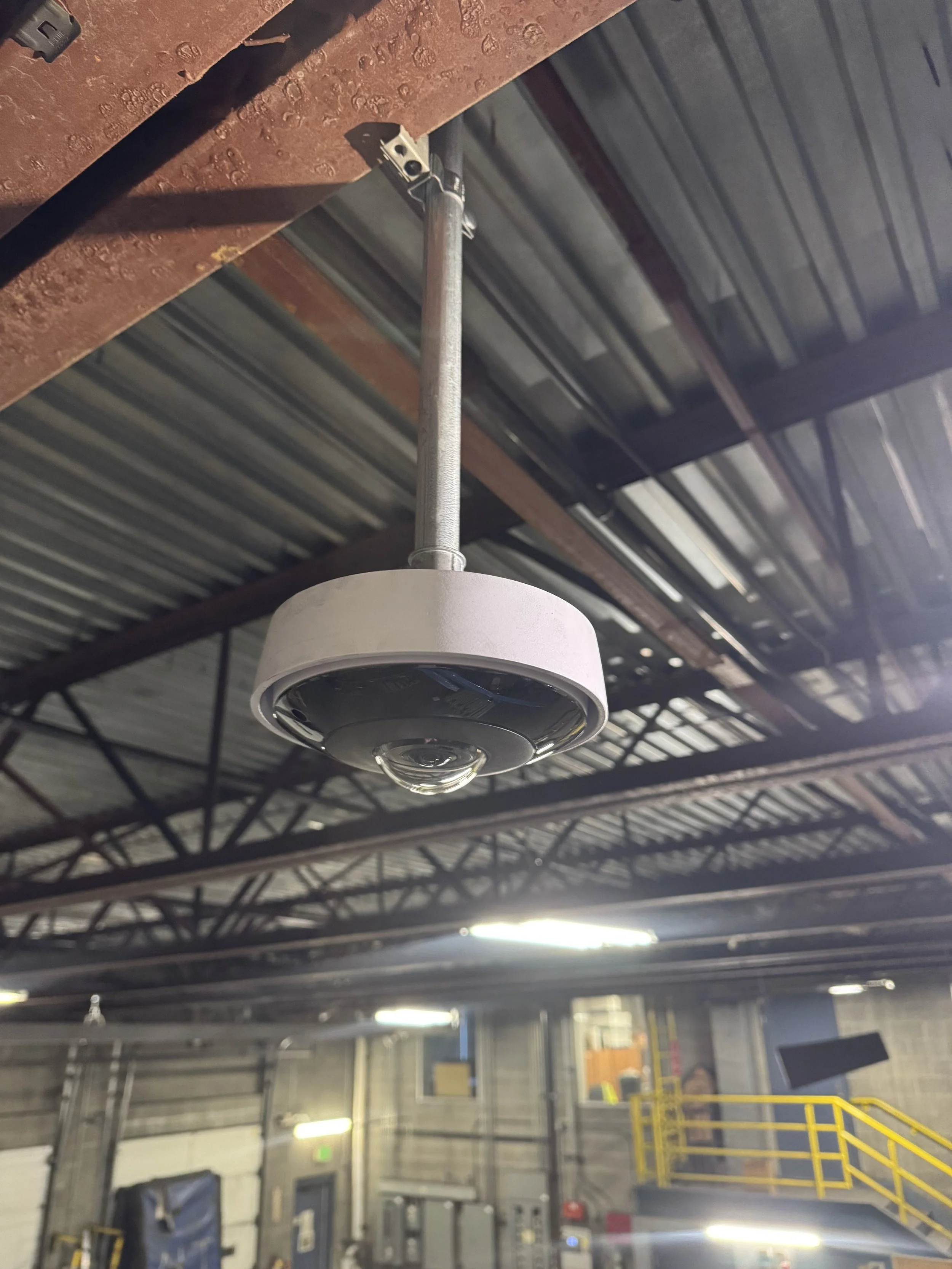 Close-up of a ceiling-mounted Verkada 360º security camera inside an industrial warehouse building with metal beams and lighting.