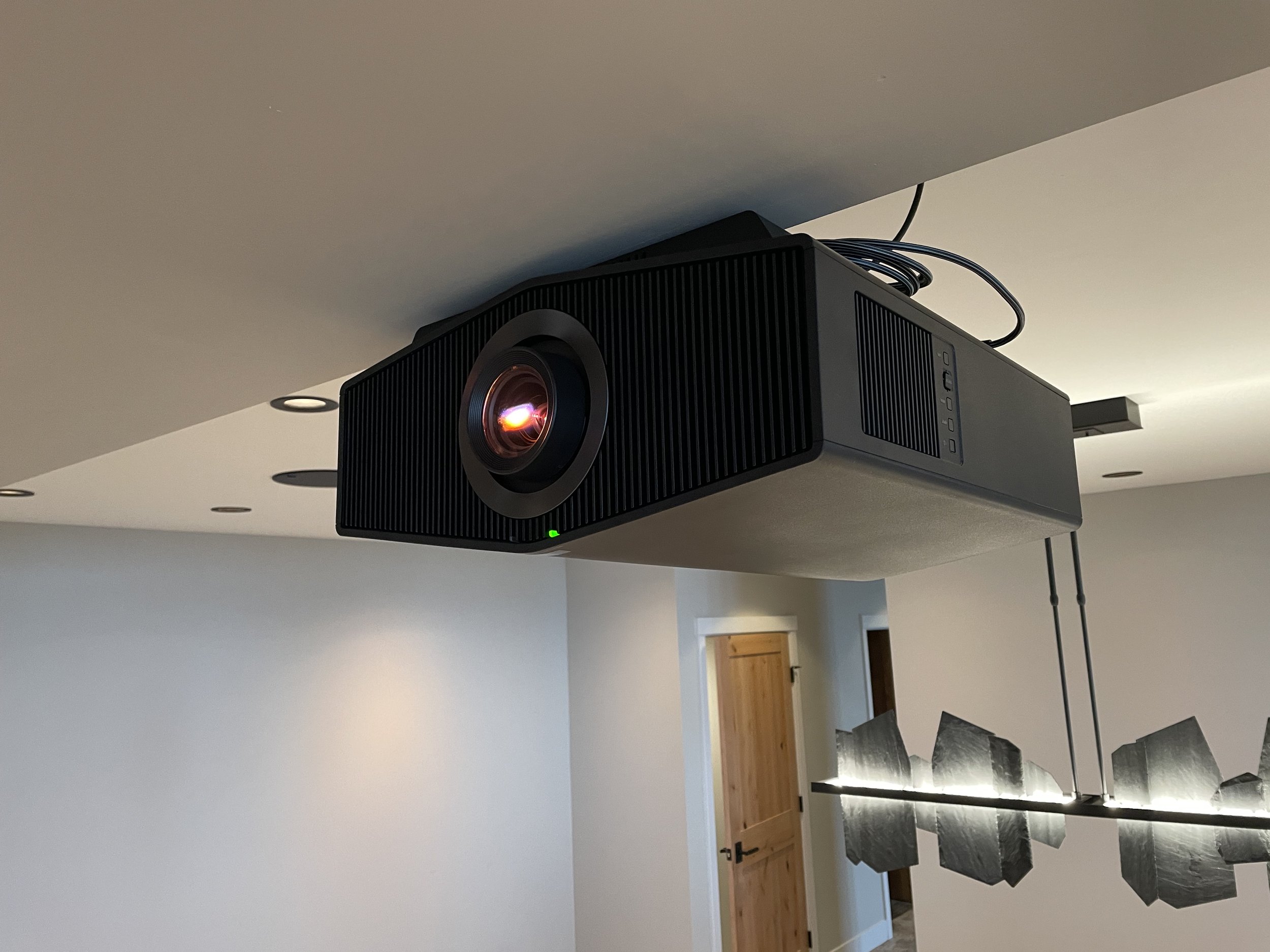 A Black Sony 4K HDR Laser projector mounted on ceiling in modern living room with decorative metal wall art and wooden door in background.