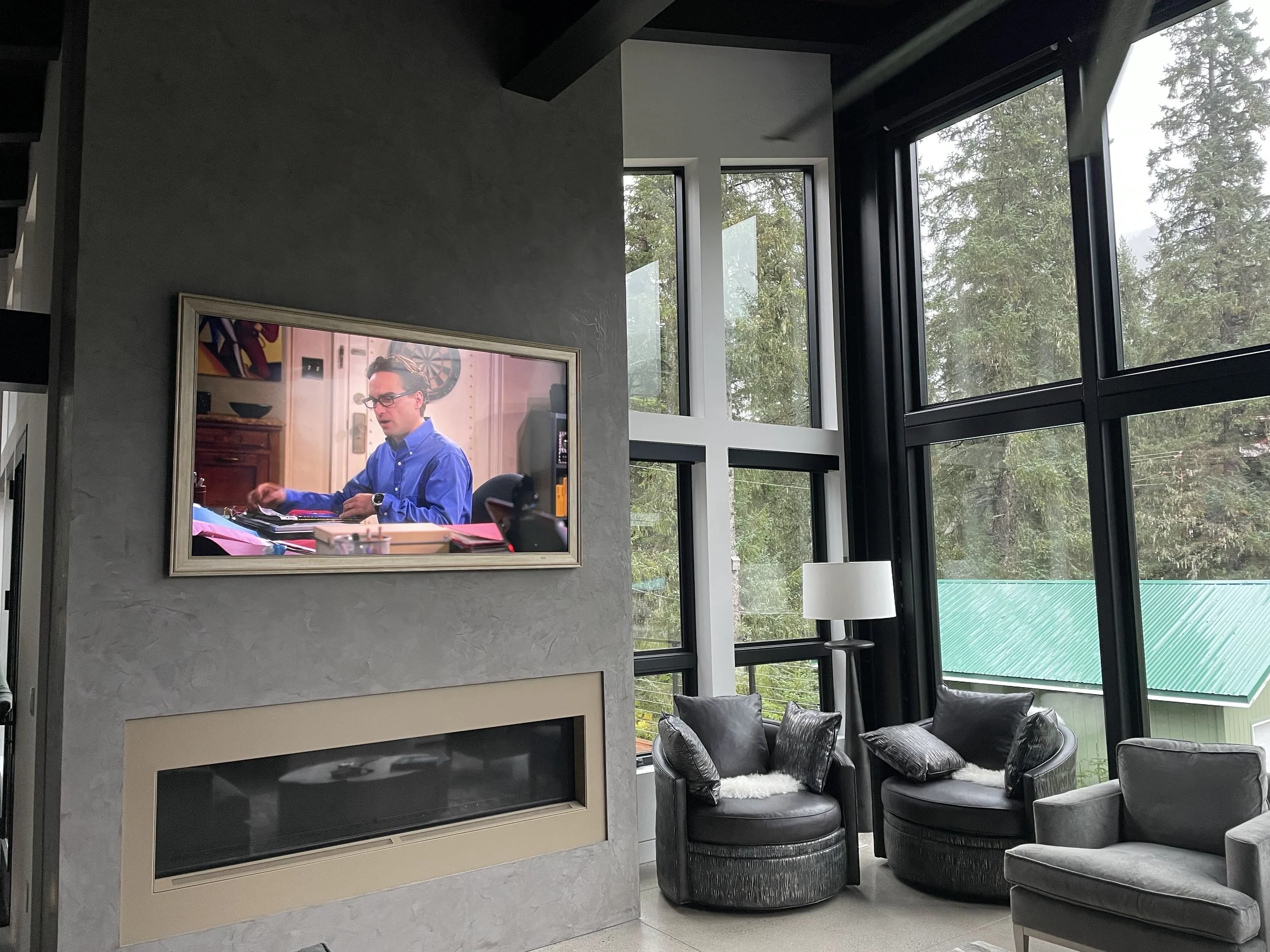Modern living room with large black-framed windows, a wall-mounted TV showing a man in a blue shirt, a fireplace below the TV, and black and gray chairs with cushions.