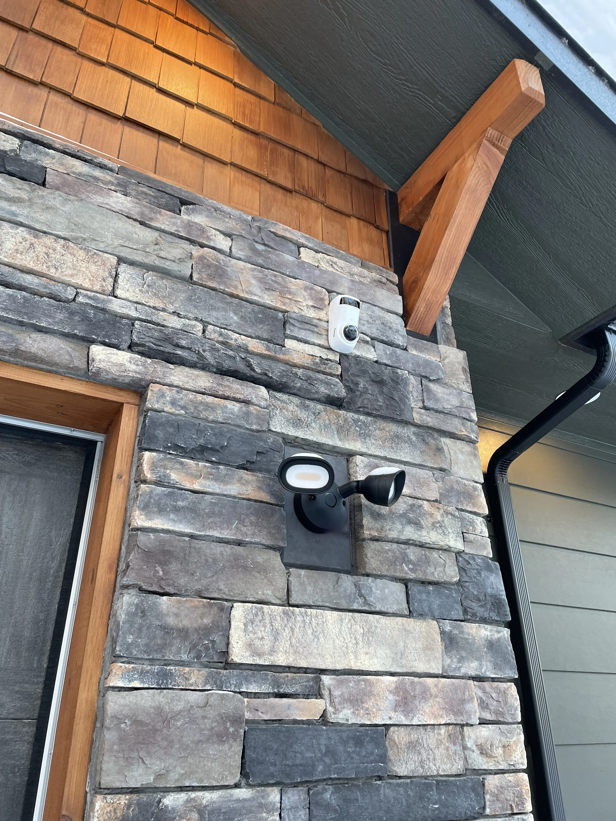 Close-up of an exterior wall with stone veneer, wood trim, a security camera, and a modern outdoor light fixture.