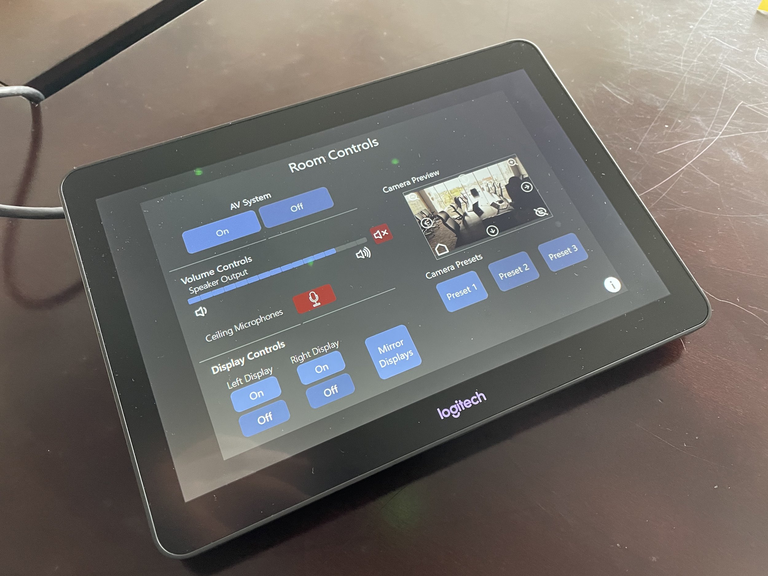 A Logitech touchscreen control panel displaying room controls such as AV system on/off, volume slider, ceiling microphone status, display controls, camera preview, camera presets, and mirror displays, placed on a dark wooden surface.