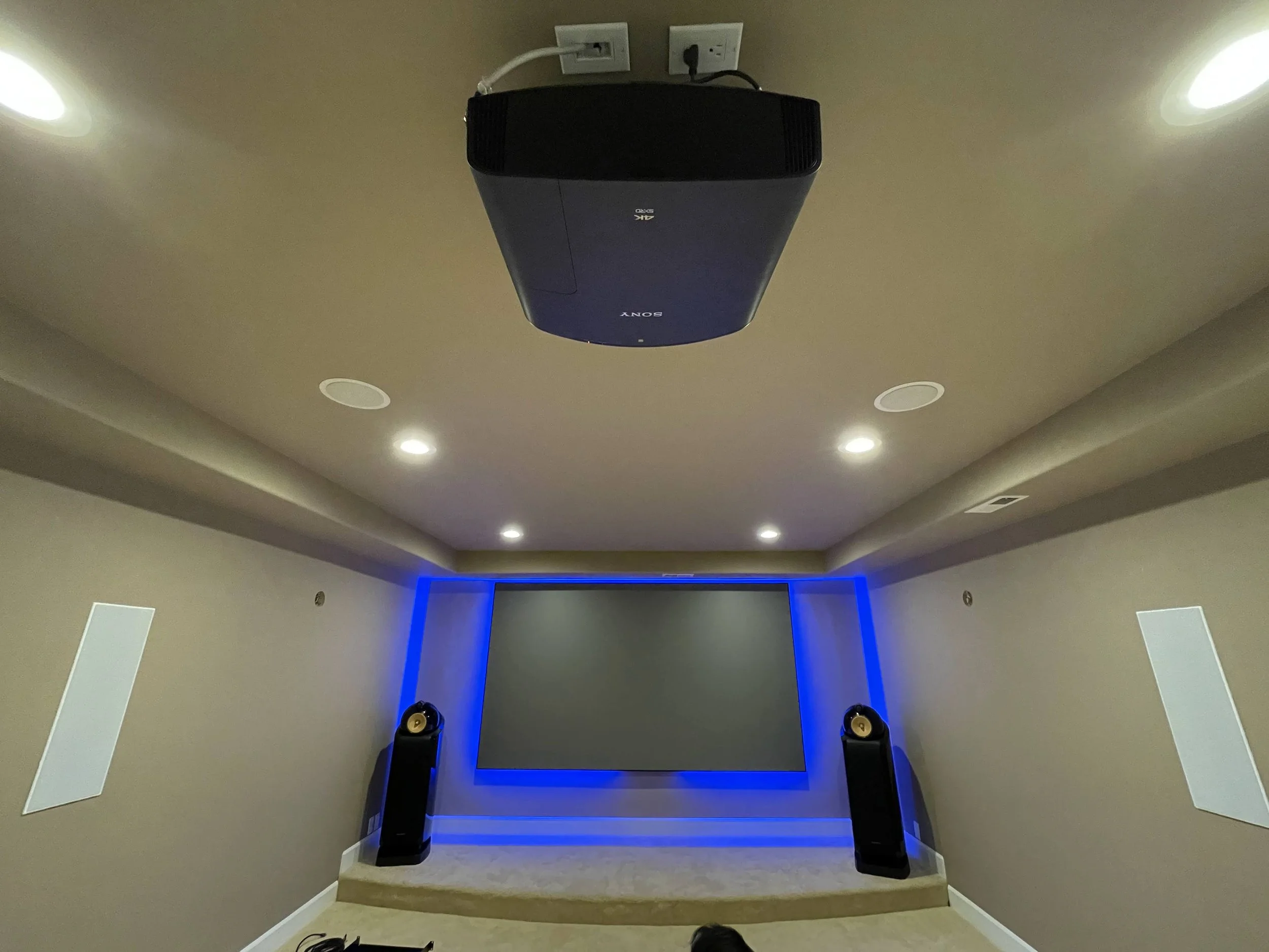 Home theater room with large Screen Innovations projection screen and Sony 4K HDR Laser Projector, blue accent backlighting, two Bowers & Wilkins tall black speakers, and recessed ceiling lights.