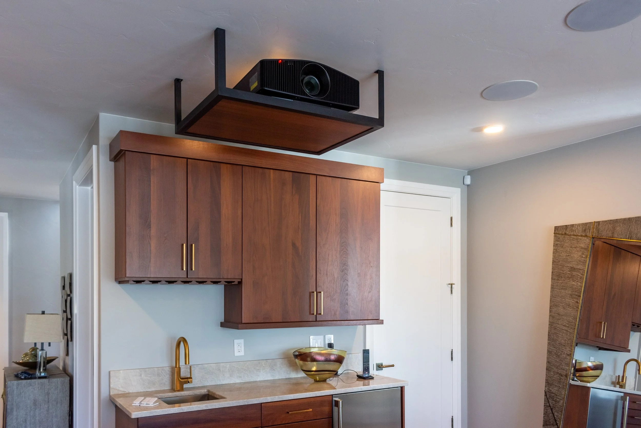 A room with a ceiling-mounted projector, a wooden cabinet, a sink with a gold faucet, and a large colorful bowl on the countertop, with a mirror and a white door in the background.
