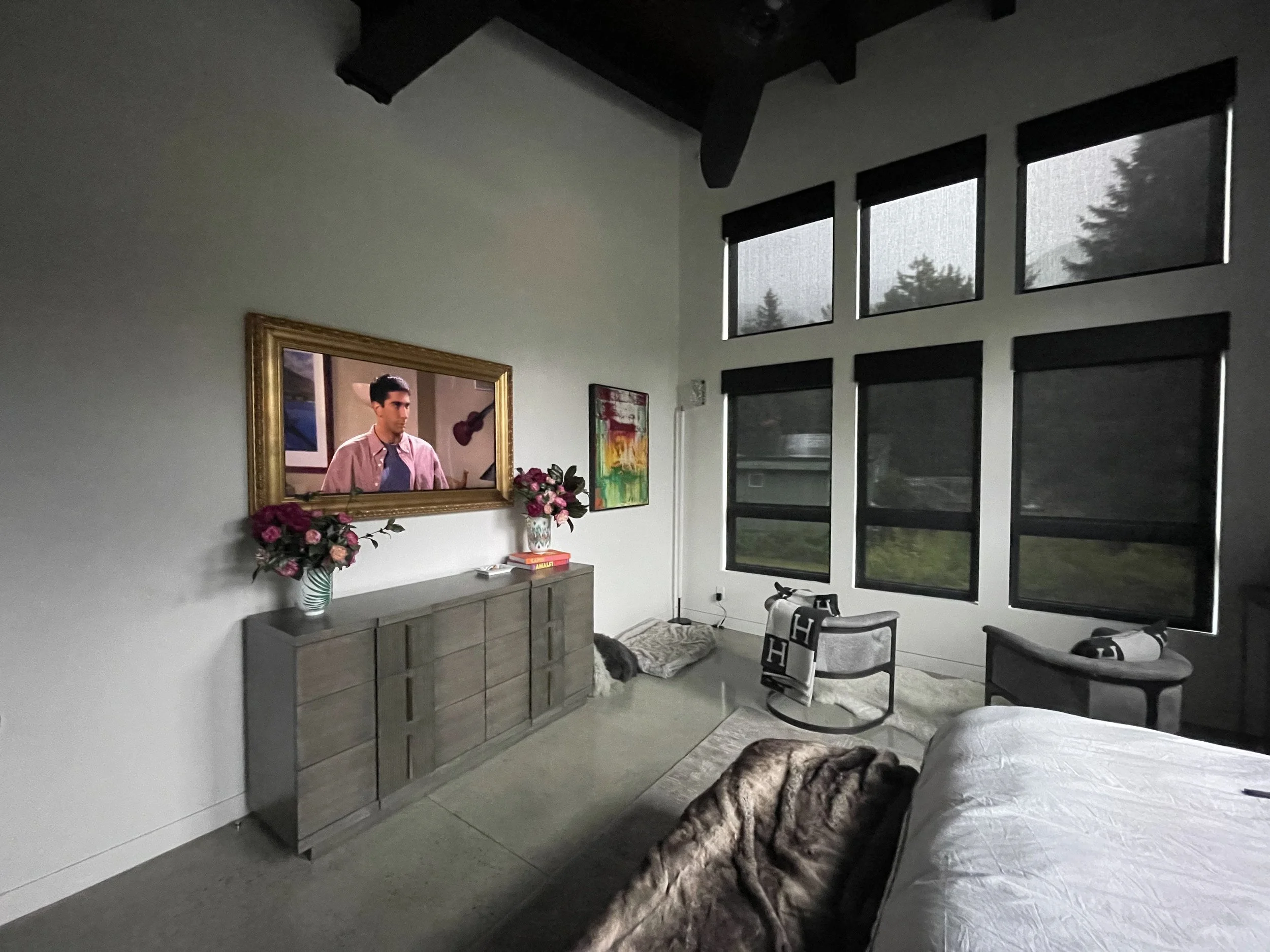 A bedroom with a bed, two chairs, a dresser with flowers, and artwork, large windows with black shades, and a TV showing a man.