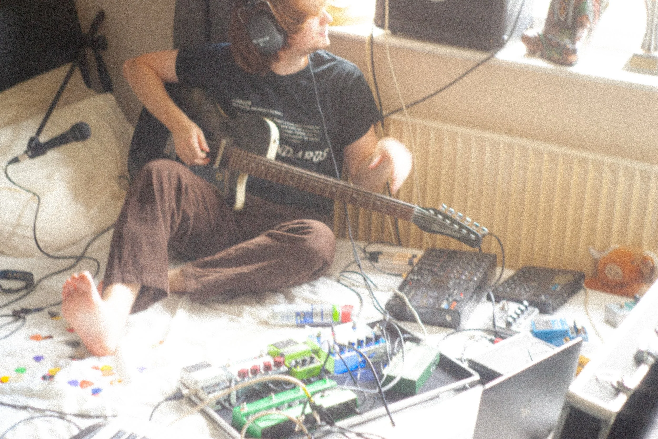 Jamie Haddington of Contact Buzz sitting on a bed, playing electric guitar amidst various music equipment like a 4-track tape recorder, guitar pedals, and electronic devices, with a window nearby and a radiator on the wall.