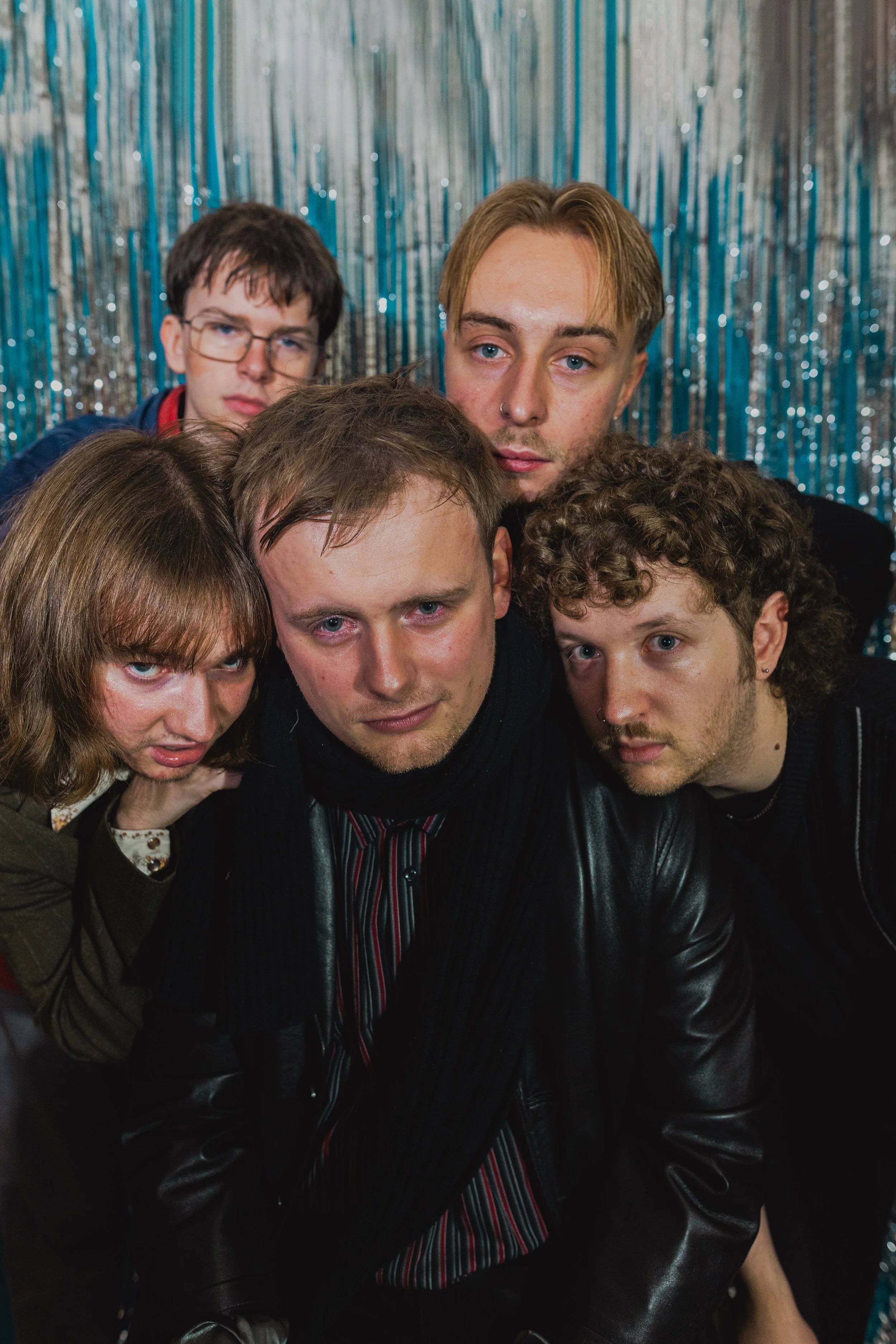 Leeds band Mince posing closely together against a sparkly blue curtain background, some leaning in and looking directly at the camera.