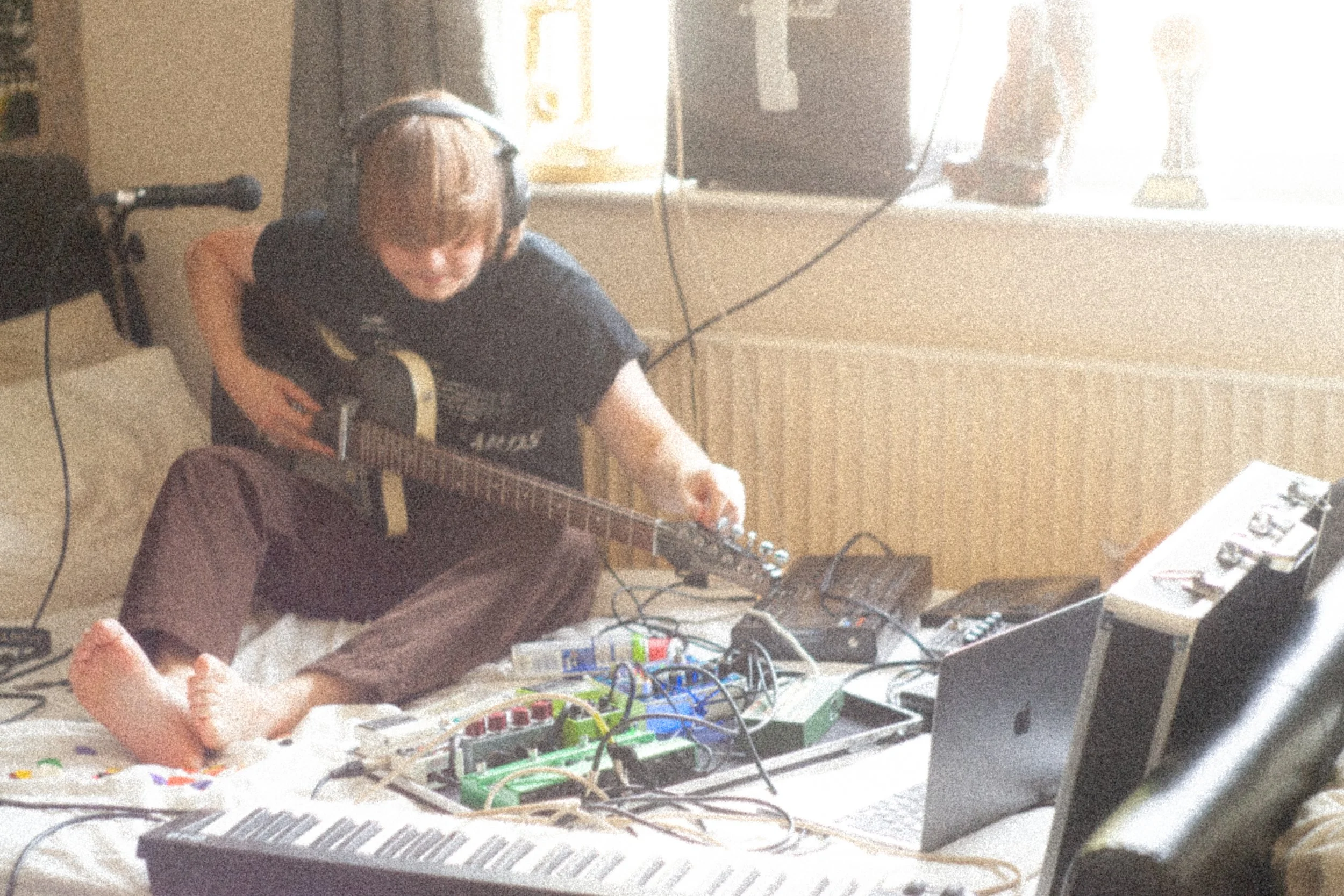 Jamie Haddington of Contact Buzz sitting on a bed playing an electric guitar, surrounded by electronic music equipment including a laptop, guitar pedals, a 4-track tape recorder and a keyboard, in a room with a radiator and a window.