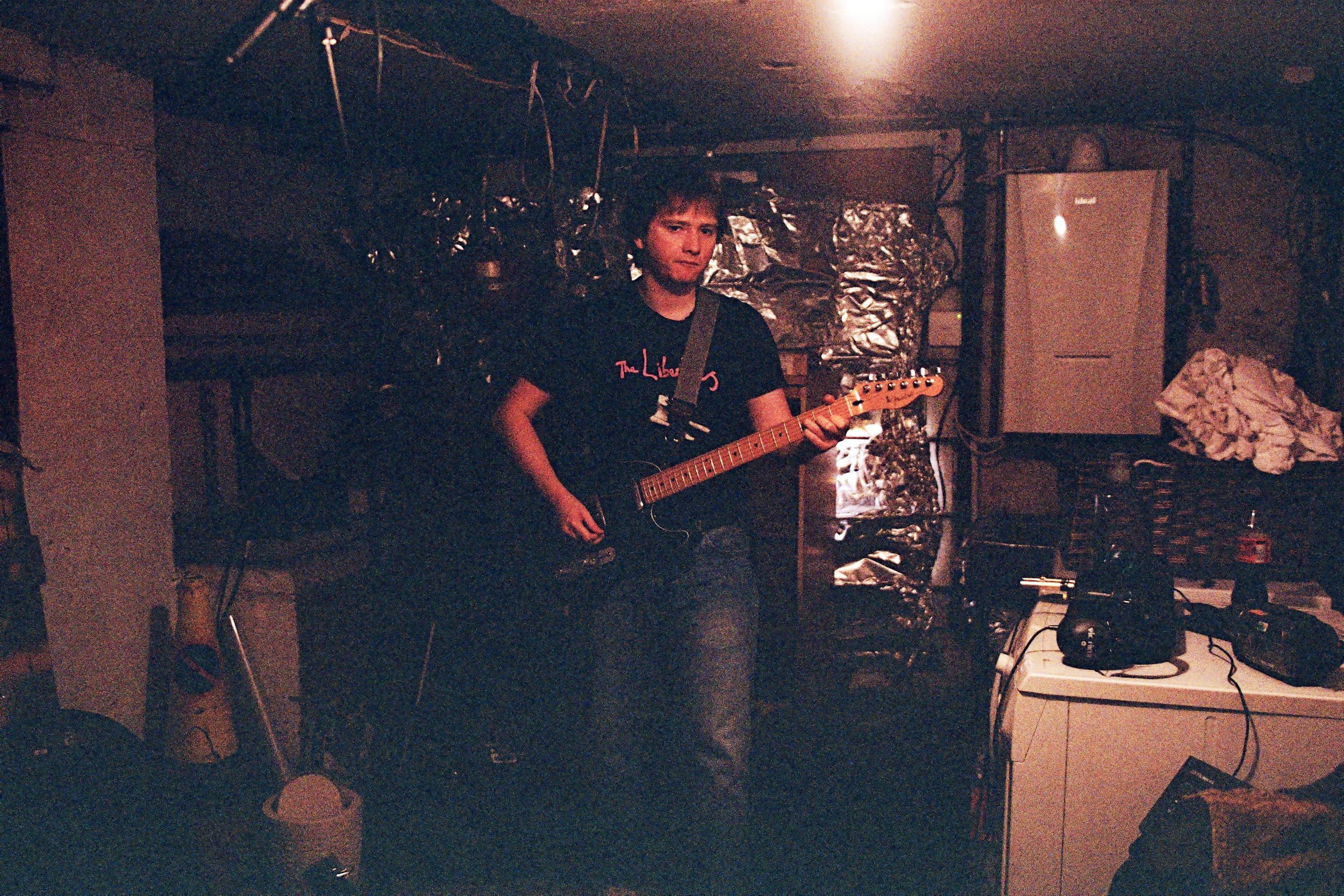 Jamie Haddington of Contact Buzz playing an electric guitar in a dimly lit basement, with various his 4-track recording equipment scattered around.