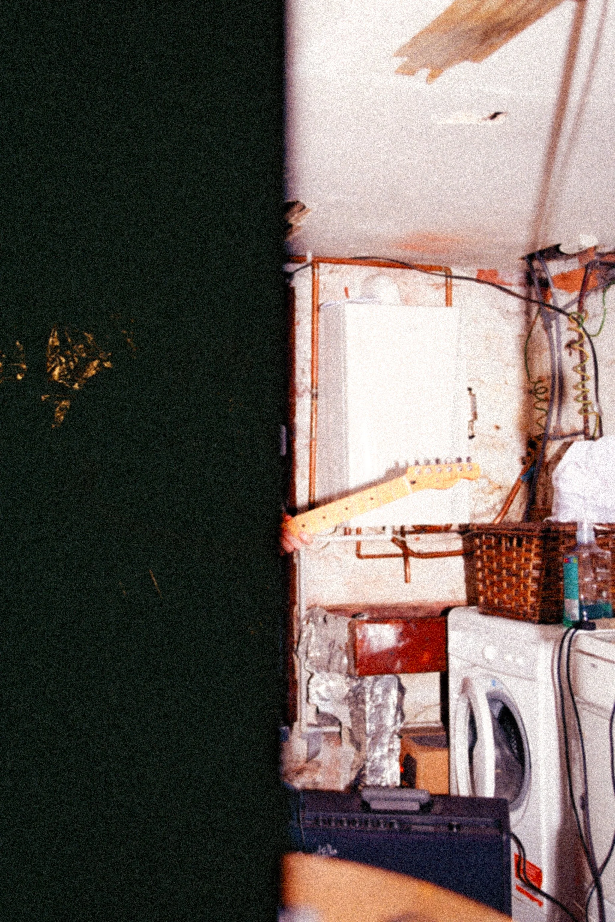 A darkened room with a partial view of a white washing machine, a wicker basket, electronic cords, and Jamie Haddington of Contact Buzz's guitar emerging from the darkness.