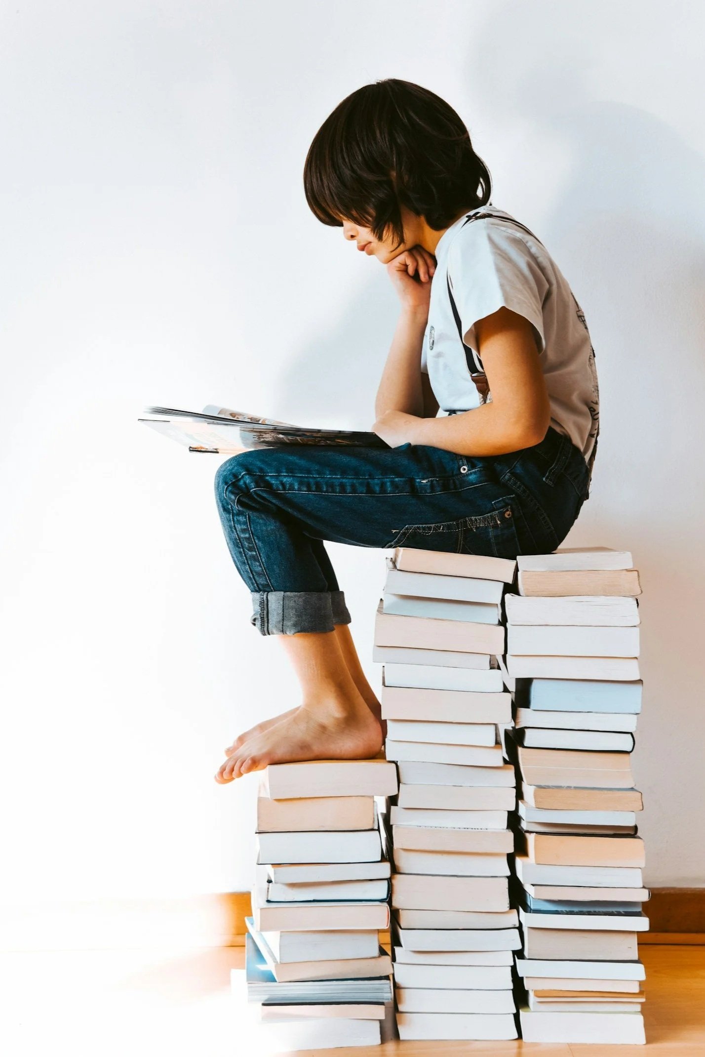 A child sits on a stack of books, reading a book, with his head resting on his hand, in a minimalist setting with a plain white wall background.