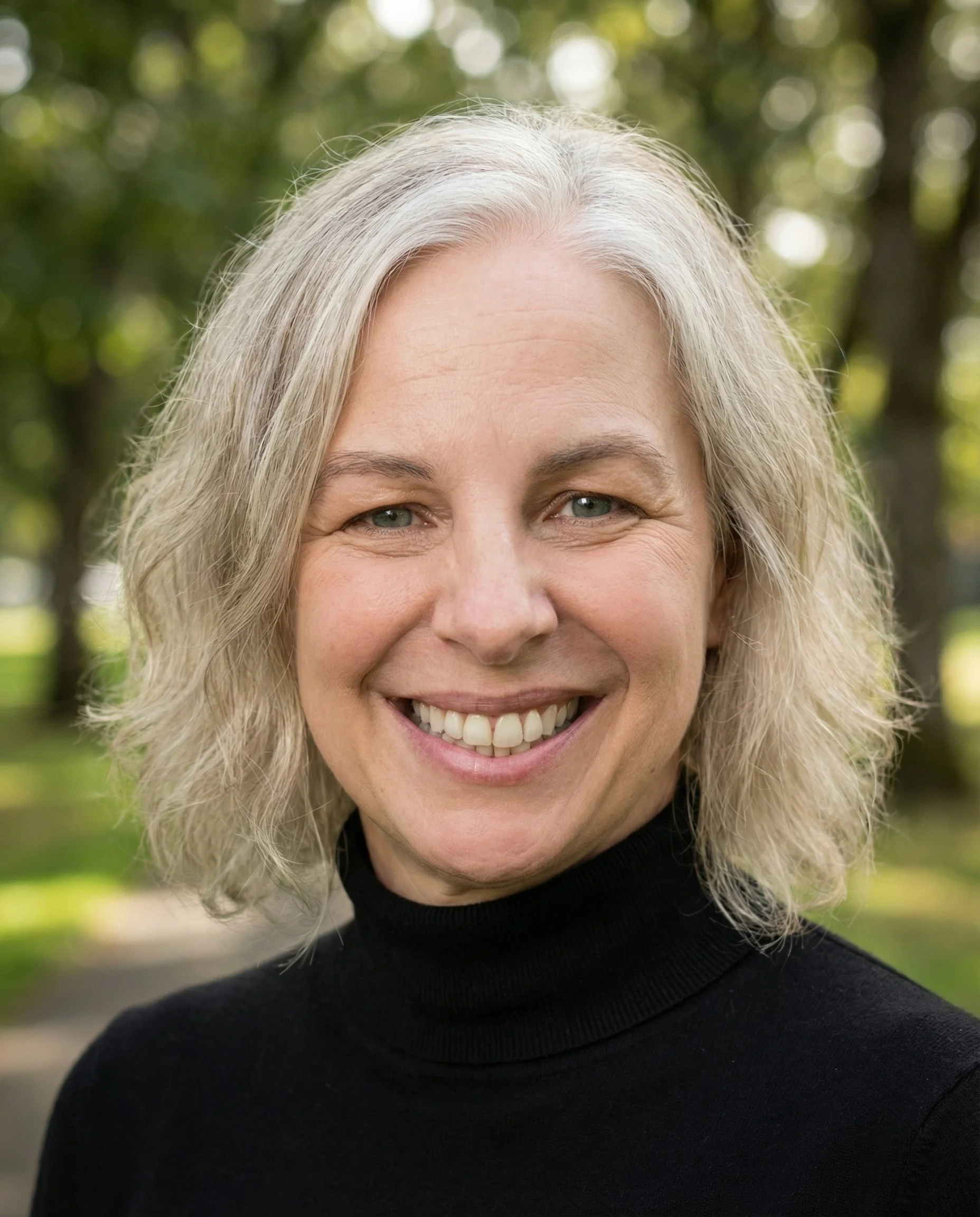 A woman with blonde hair smiling outdoors, wearing a black turtleneck, with a blurred green background.