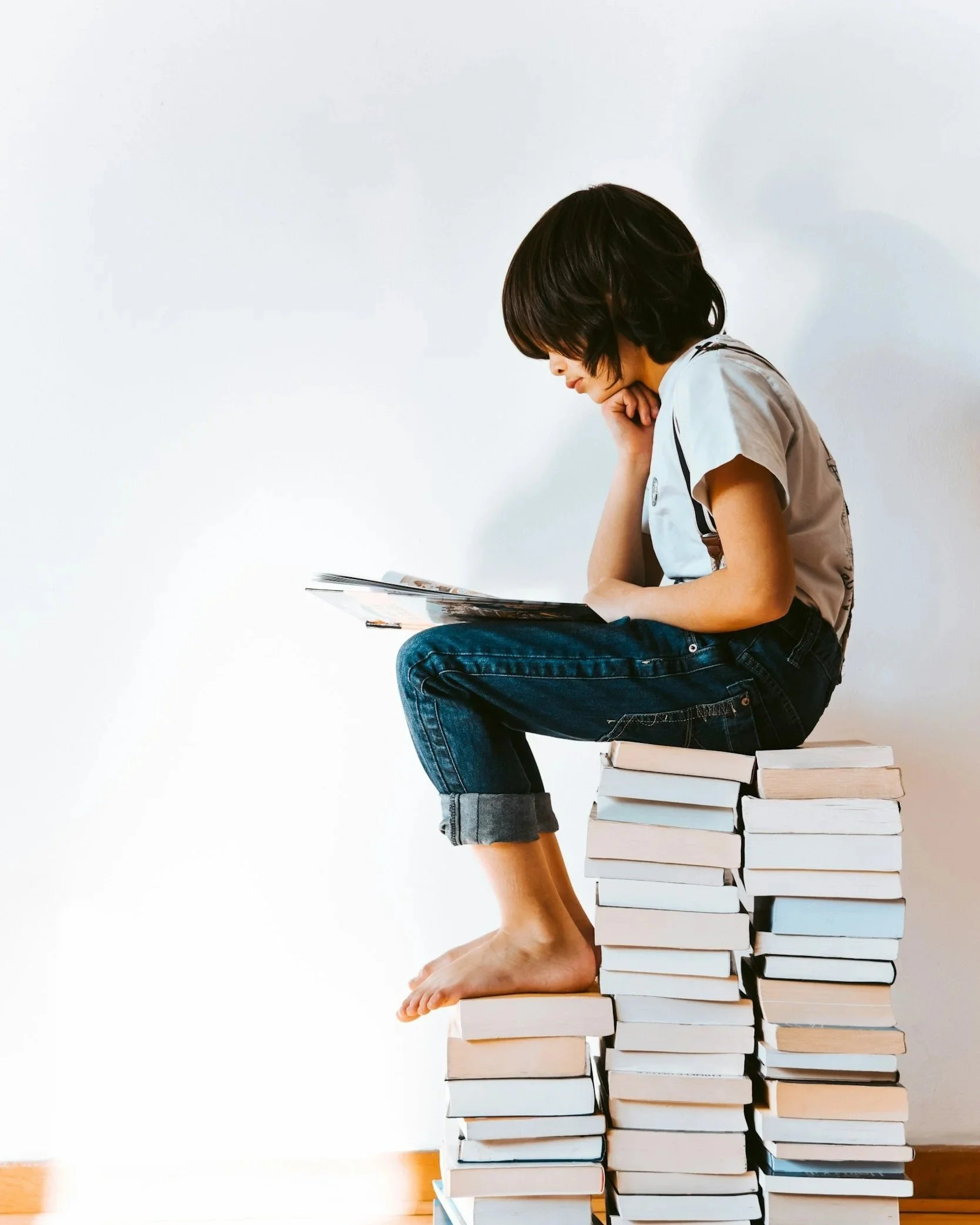 A child sits on a stack of books, reading a book, with his head resting on his hand, in a minimalist setting with a plain white wall background.