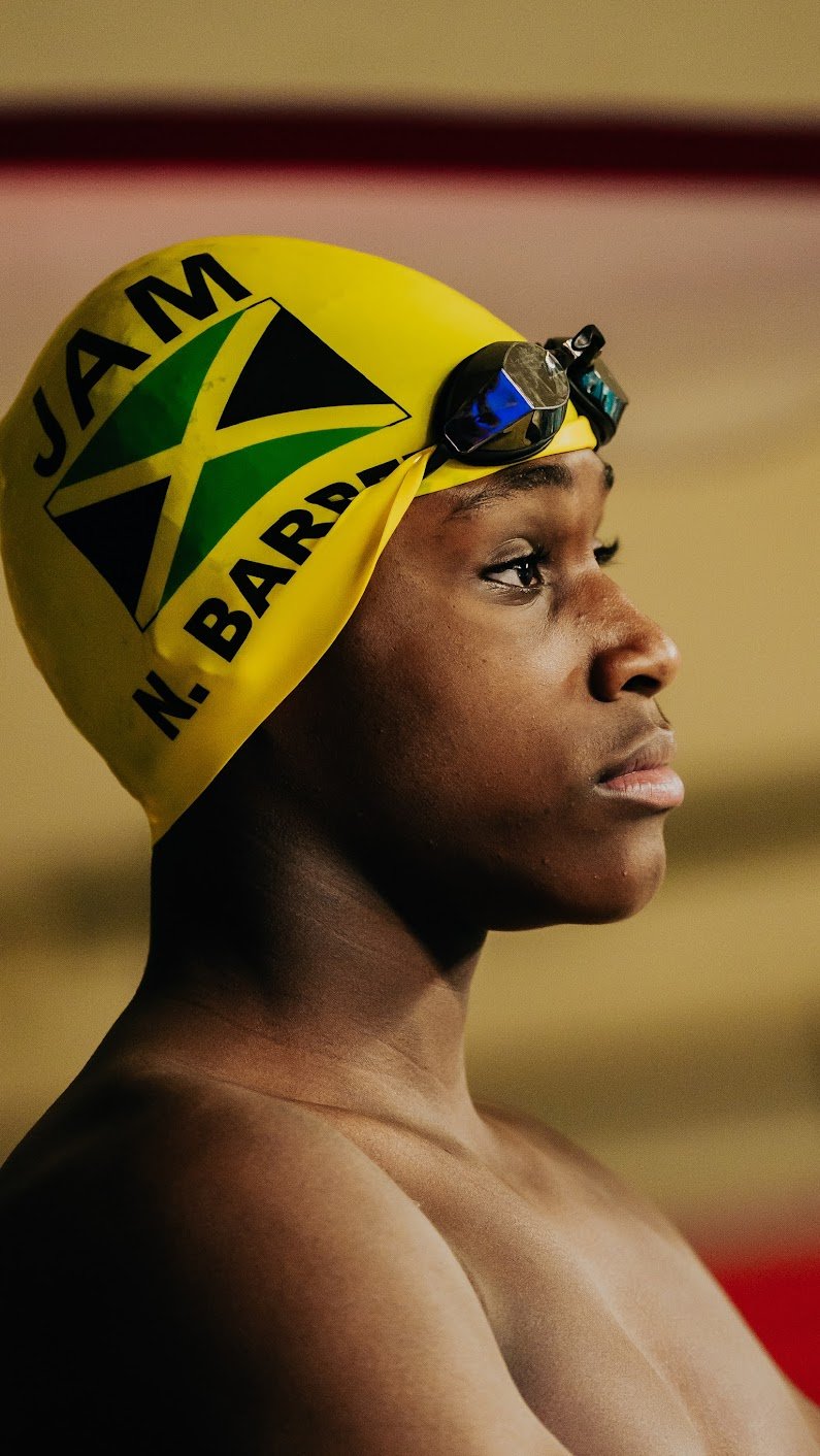 Profile of Noland wearing a yellow swim cap with Jamaican flag and text, goggles on her forehead, looking focused.