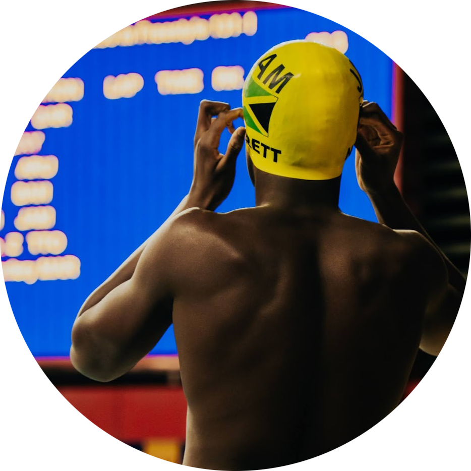 Backstage view of Noland wearing a yellow swim cap with Jamaican flag and text, adjusting the cap in front of a blue scoreboard.