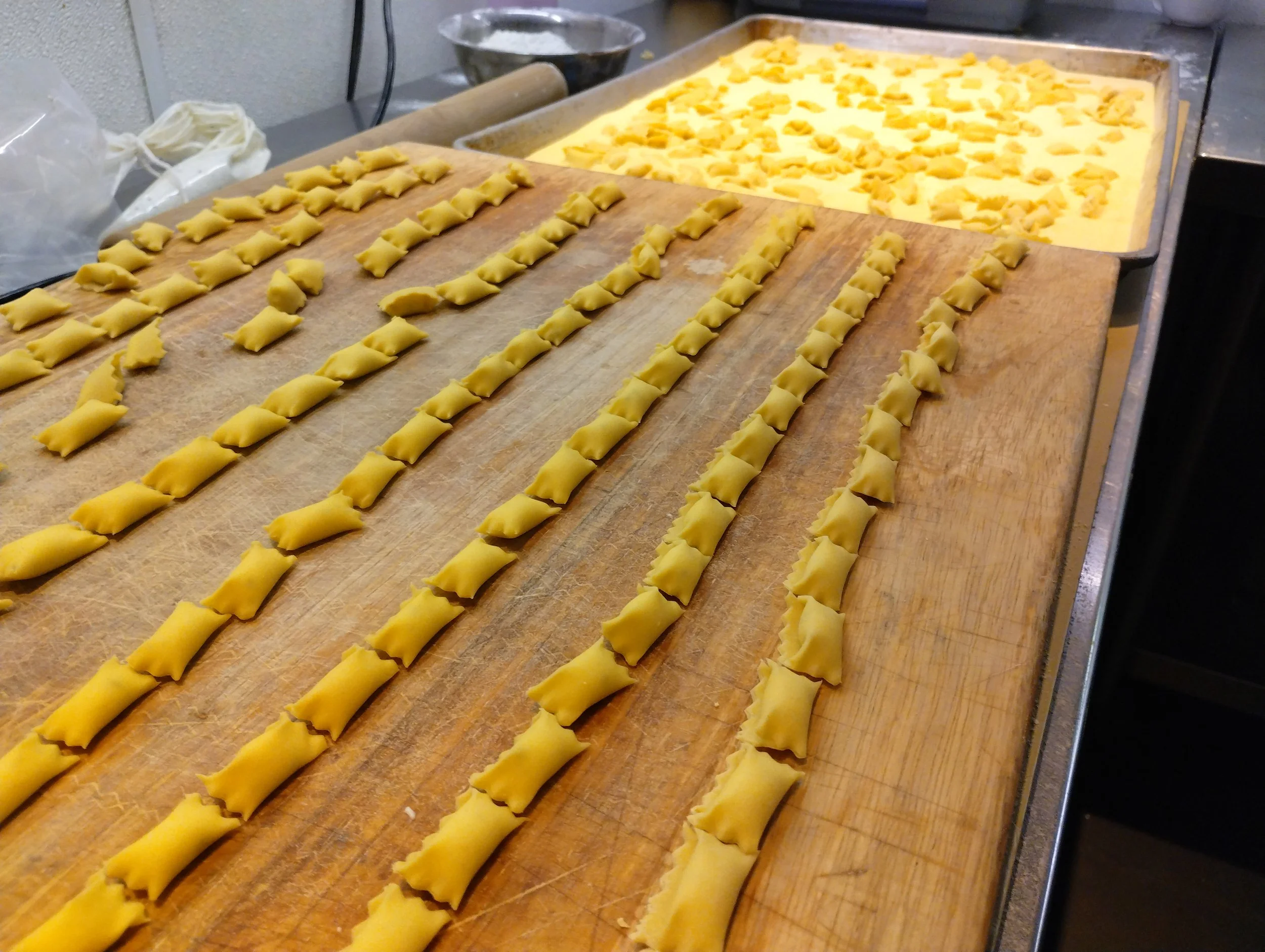 Uncooked agnolotti pasta arranged in rows on a wooden cutting board, with more filled pasta in a tray in the background.