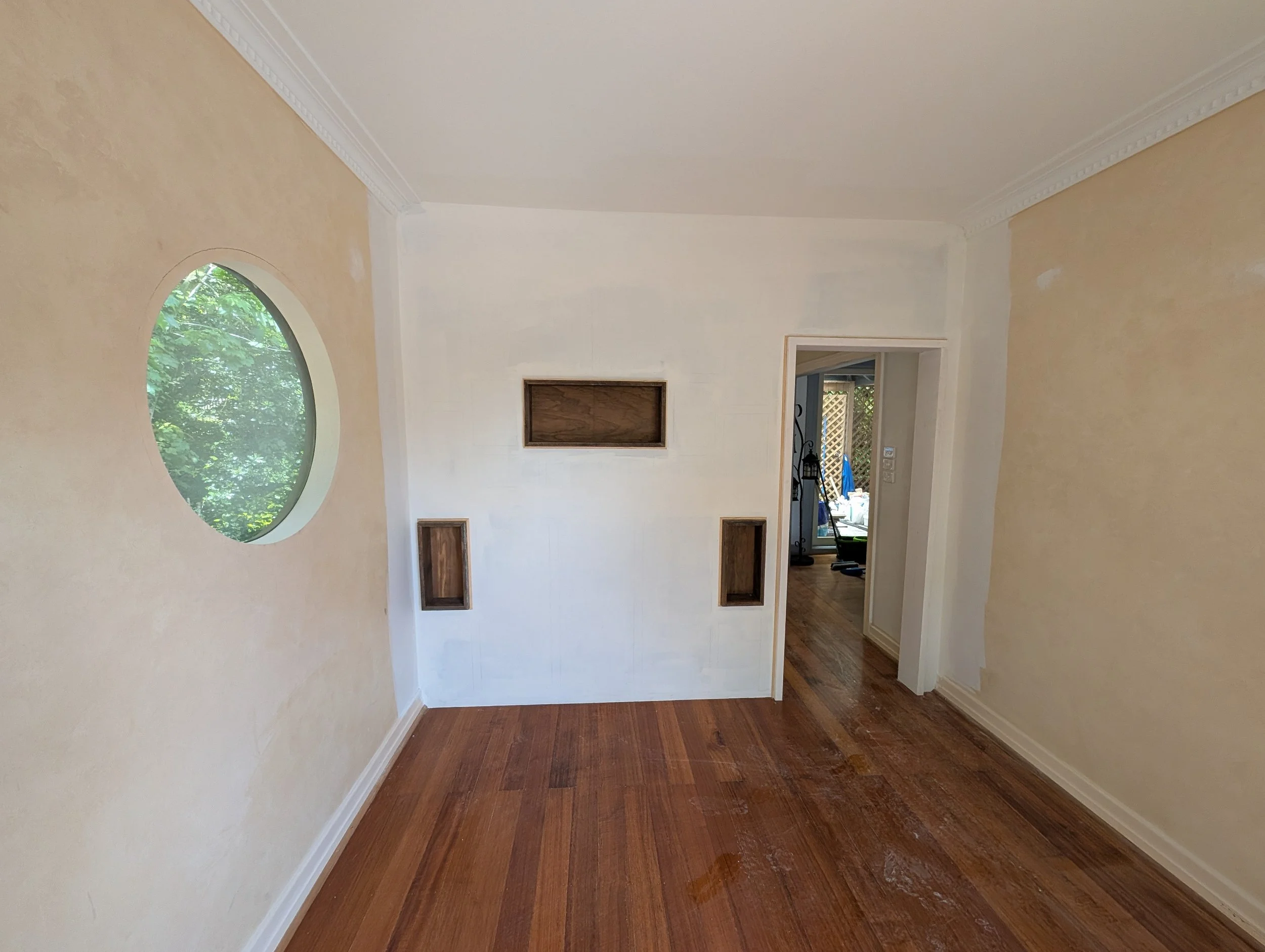 Living room with a circular window, wooden floors, and white walls with wall niches. A doorway leads to another room with belongings visible.