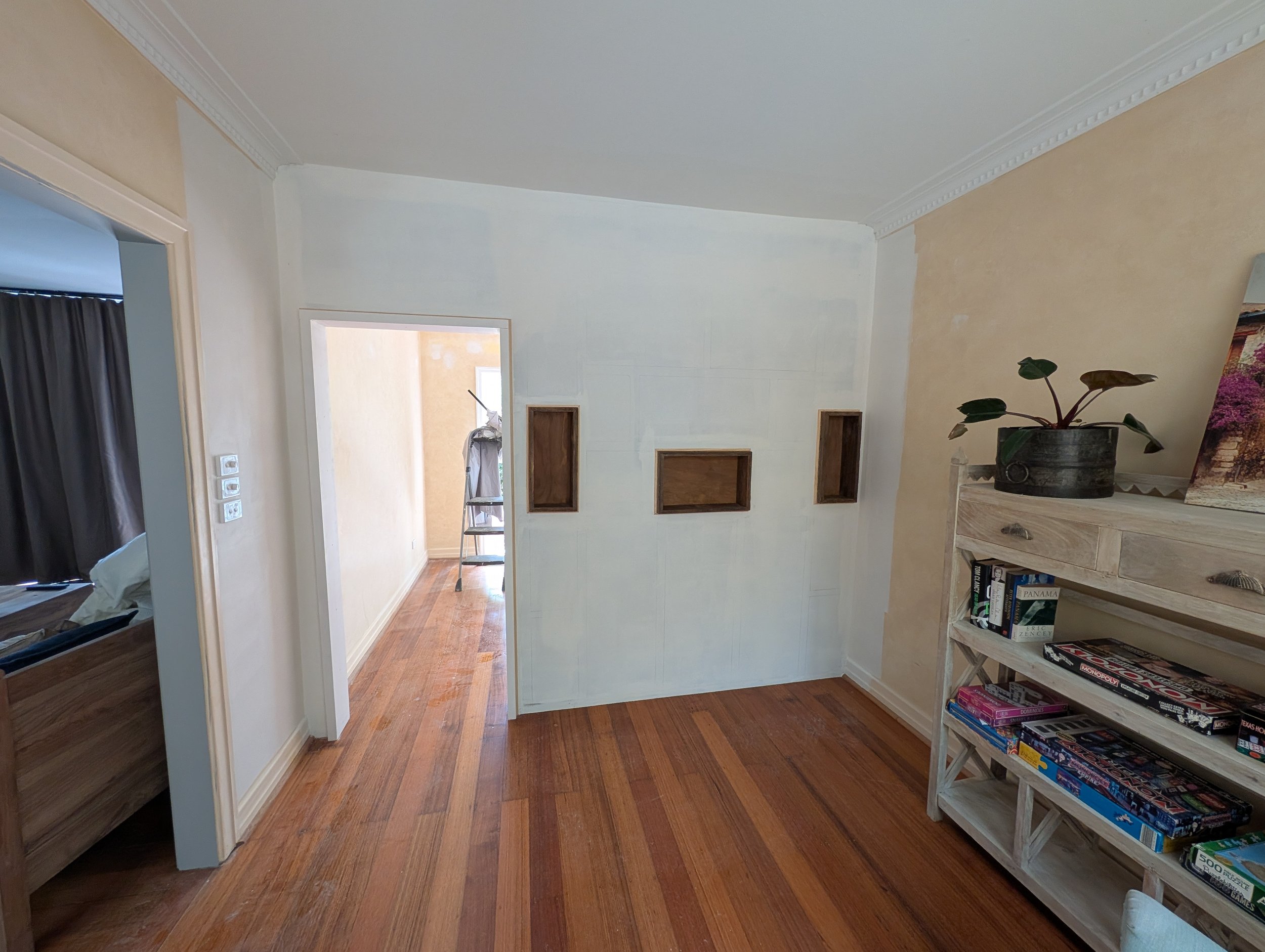 An interior view of a room with wooden flooring, a white wall with three small rectangular wooden insets, an open doorway, and a bookshelf filled with board games. A potted plant is on top of a wooden cabinet near the bookshelf.