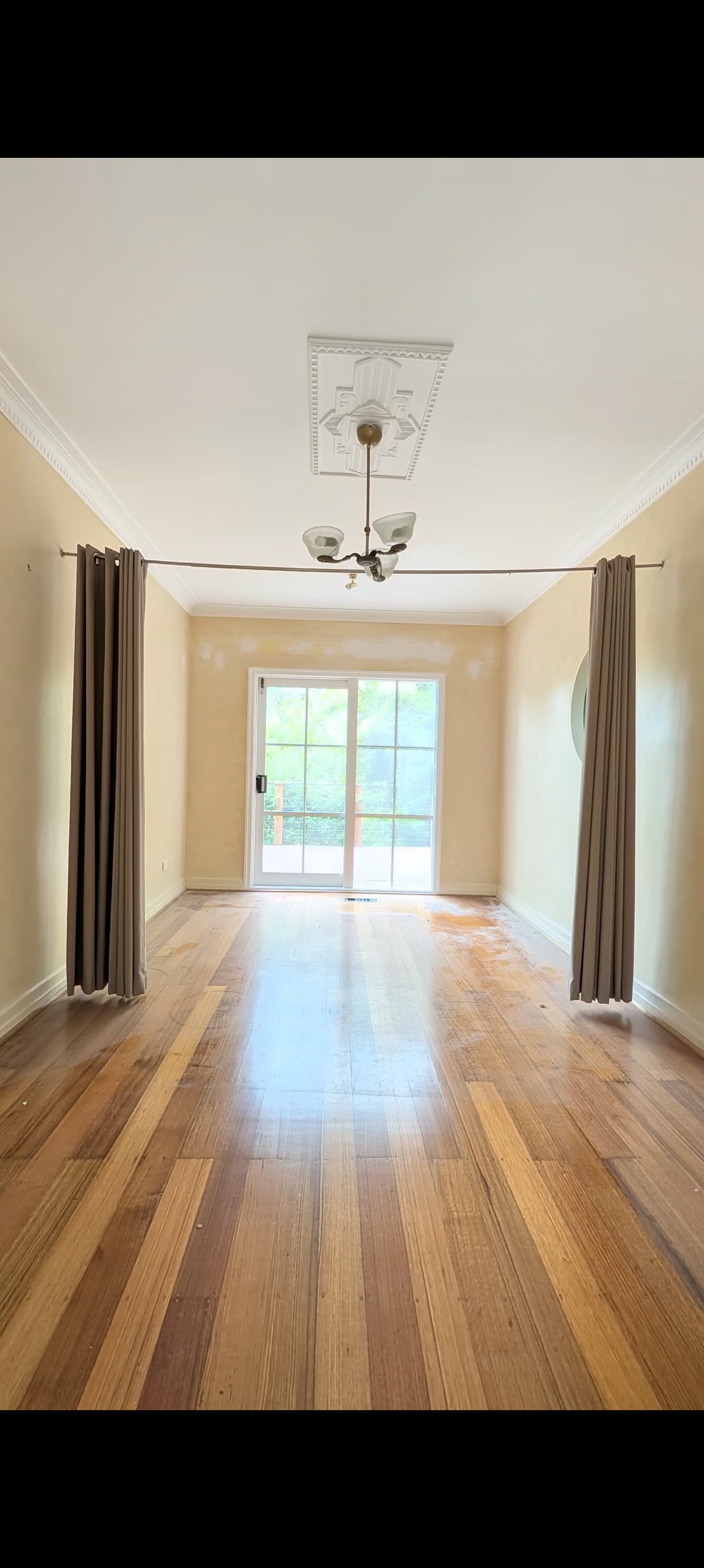 Empty room with hardwood floors, beige walls, and a sliding glass door leading outside. Brown curtains hang on a rod on either side of the door, and there is a ceiling mounted light fixture.