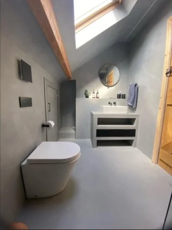 Minimalist bathroom with a sloped ceiling and skylight, gray walls, a white toilet, a black round mirror, a white vanity, and wooden accents.