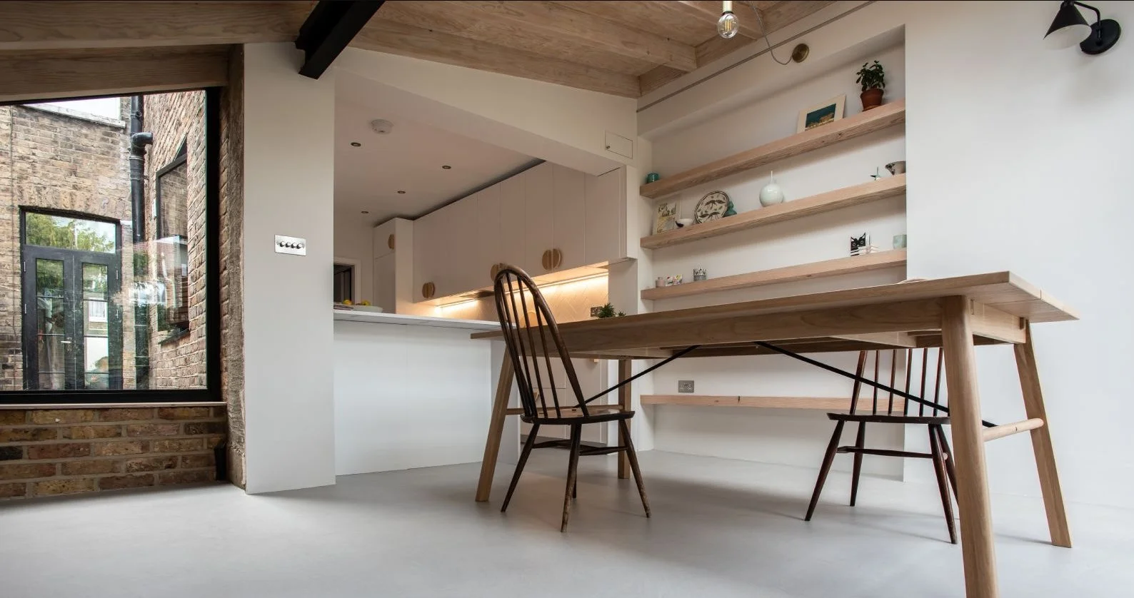 Minimalist dining area featuring a wooden table, two mid-century modern chairs, and open wooden shelves with decorative items on a white wall, next to a large window showing exposed brick exterior.