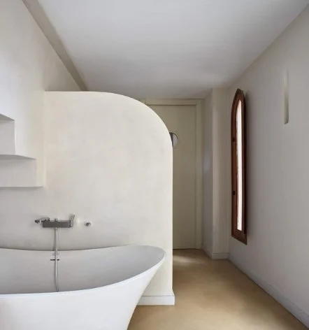 Empty interior space with a curved white bathtub, a small shelf, a door, an arched window, and simple decor.