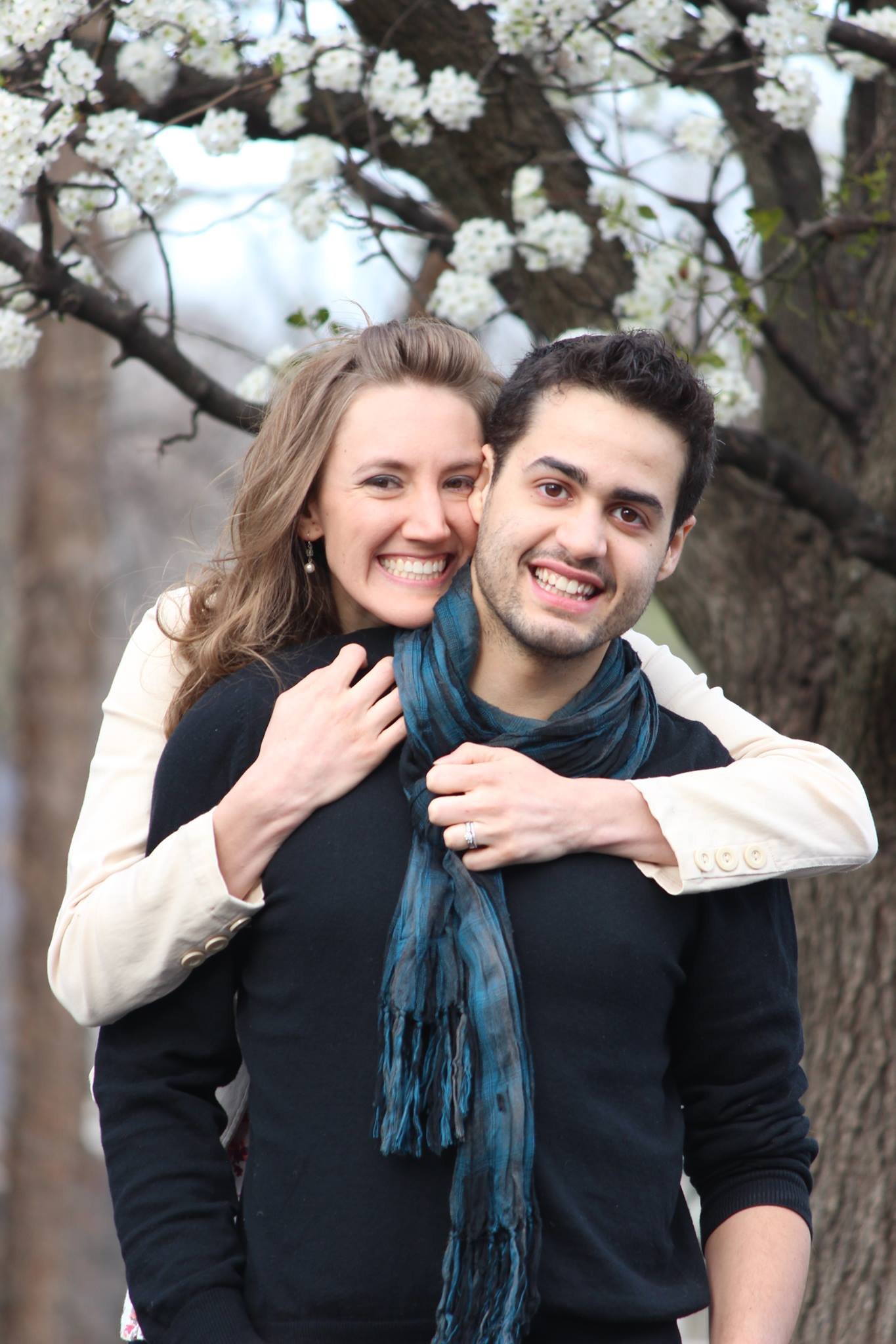 A smiling woman with wavy hair hugging a man with dark hair and a plaid scarf, outdoors near a flowering tree.