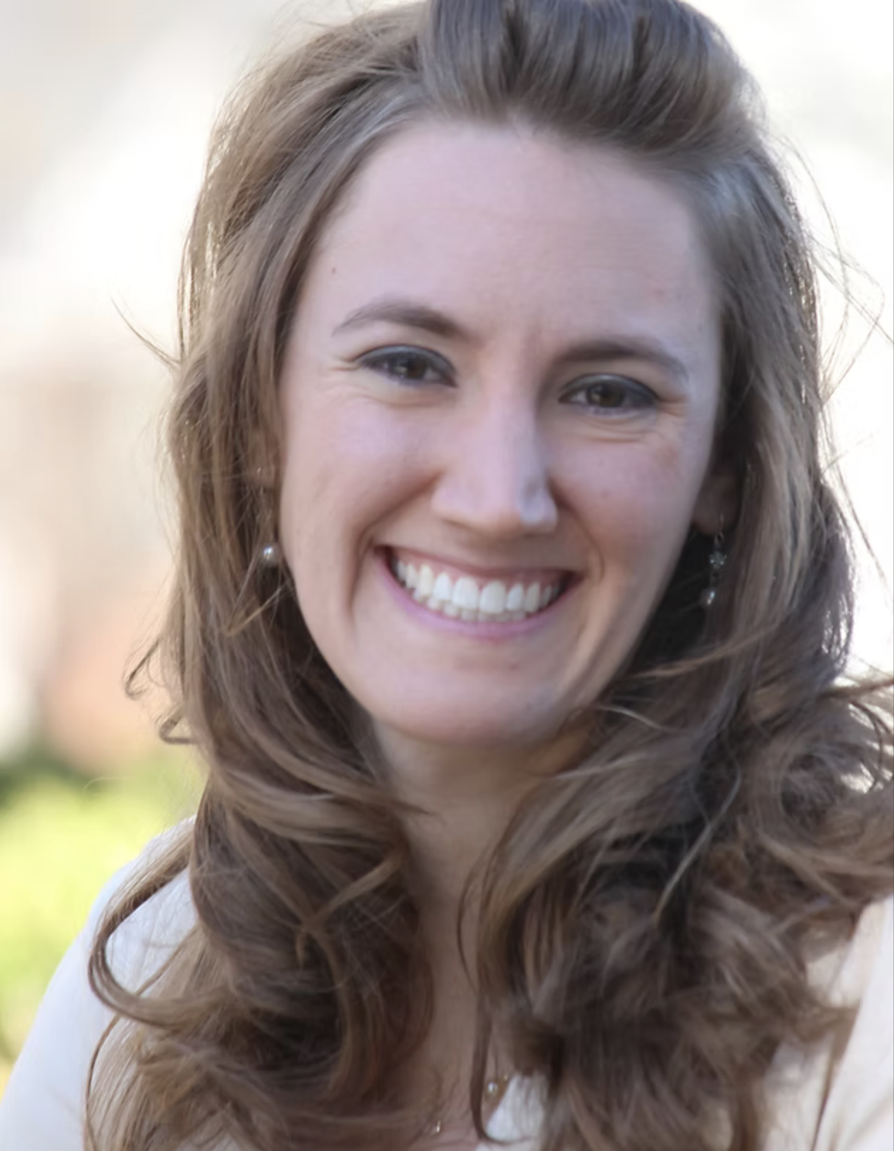 A smiling woman with long wavy brown hair outdoors, wearing pearl earrings and a light-colored top.