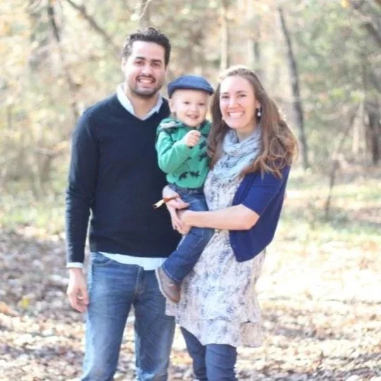 A happy family of three standing outdoors in a wooded area during autumn, with a man, a woman, and a young child smiling.